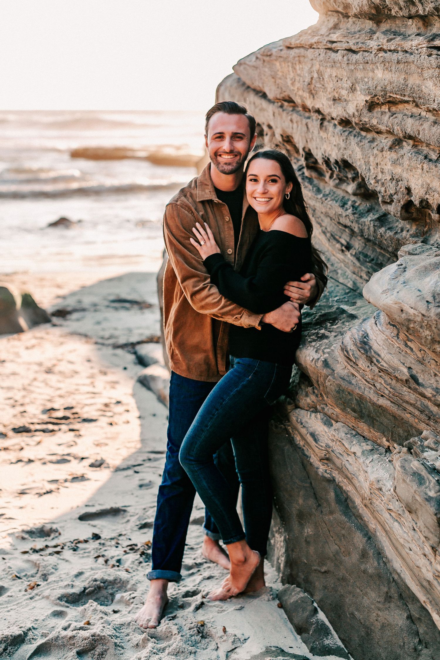 Smiling couple embracing against sandy cliffs at a beach during golden hour.