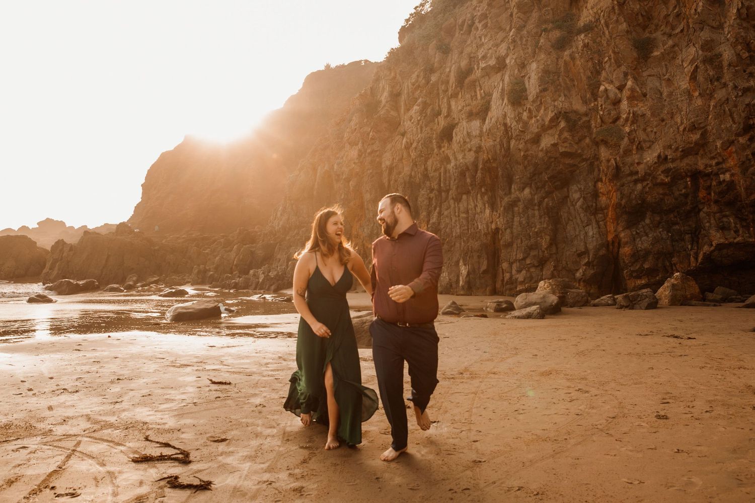 Couple walking on a sunlit beach near rocky cliffs, woman in green dress, man in dark outfit, golden hour light.