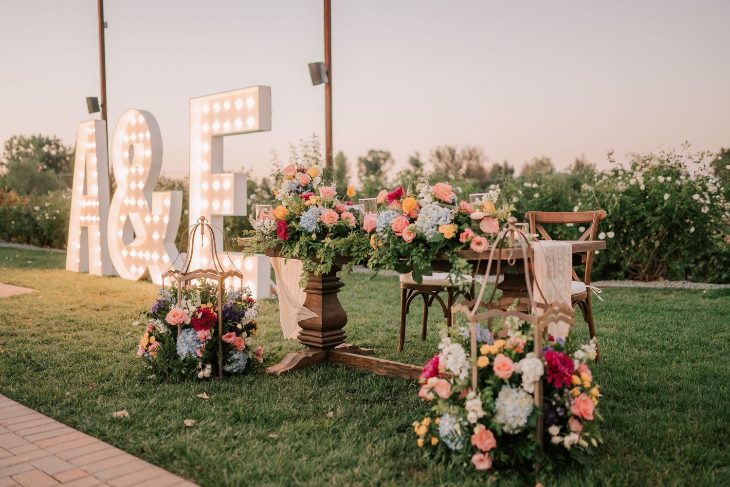 Outdoor wedding decor with marquee letters A&E, floral arrangements in pink and white blooms, and wooden chairs on green grass.
