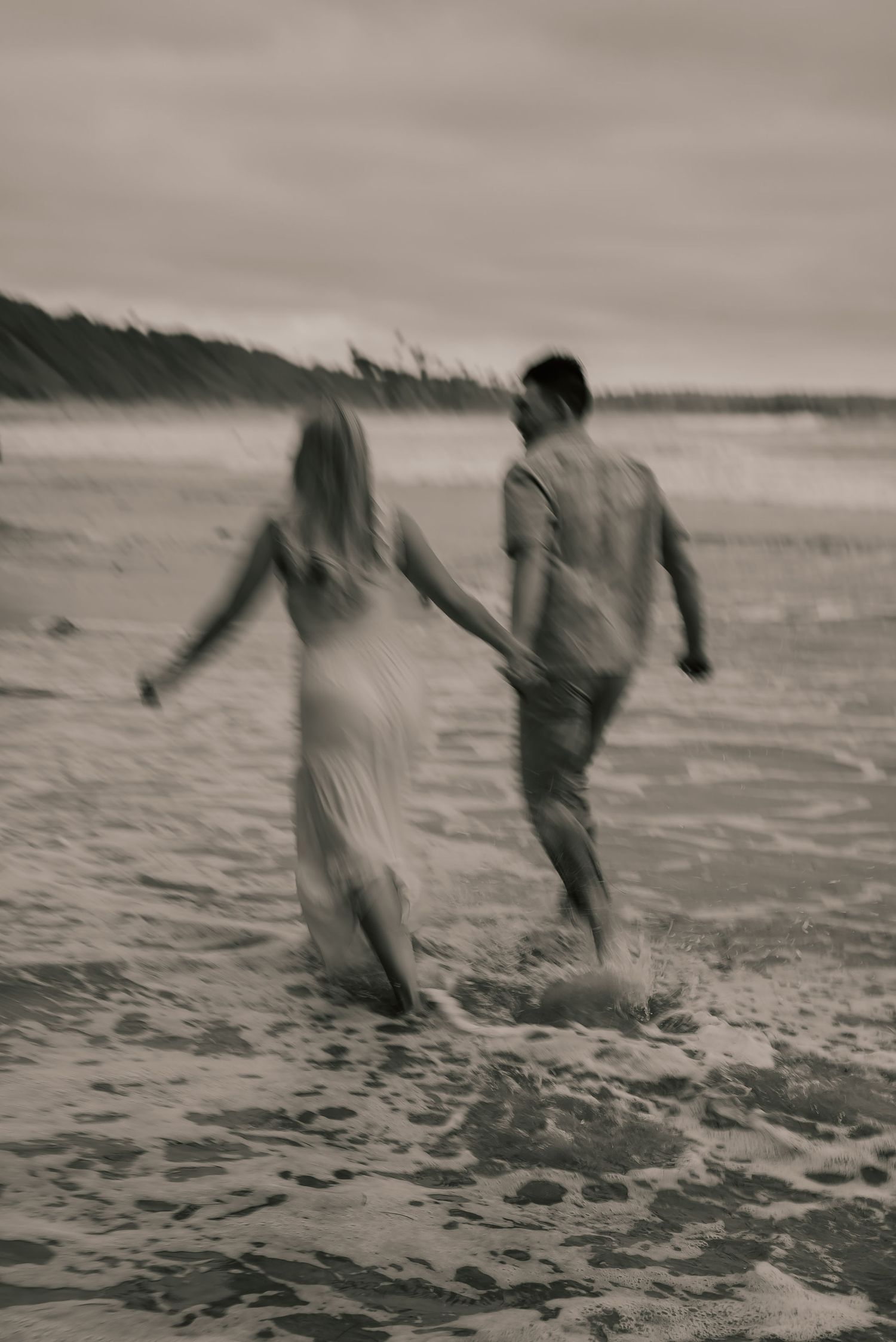 Couple holding hands running through ocean waves on beach in black and white romantic photo.