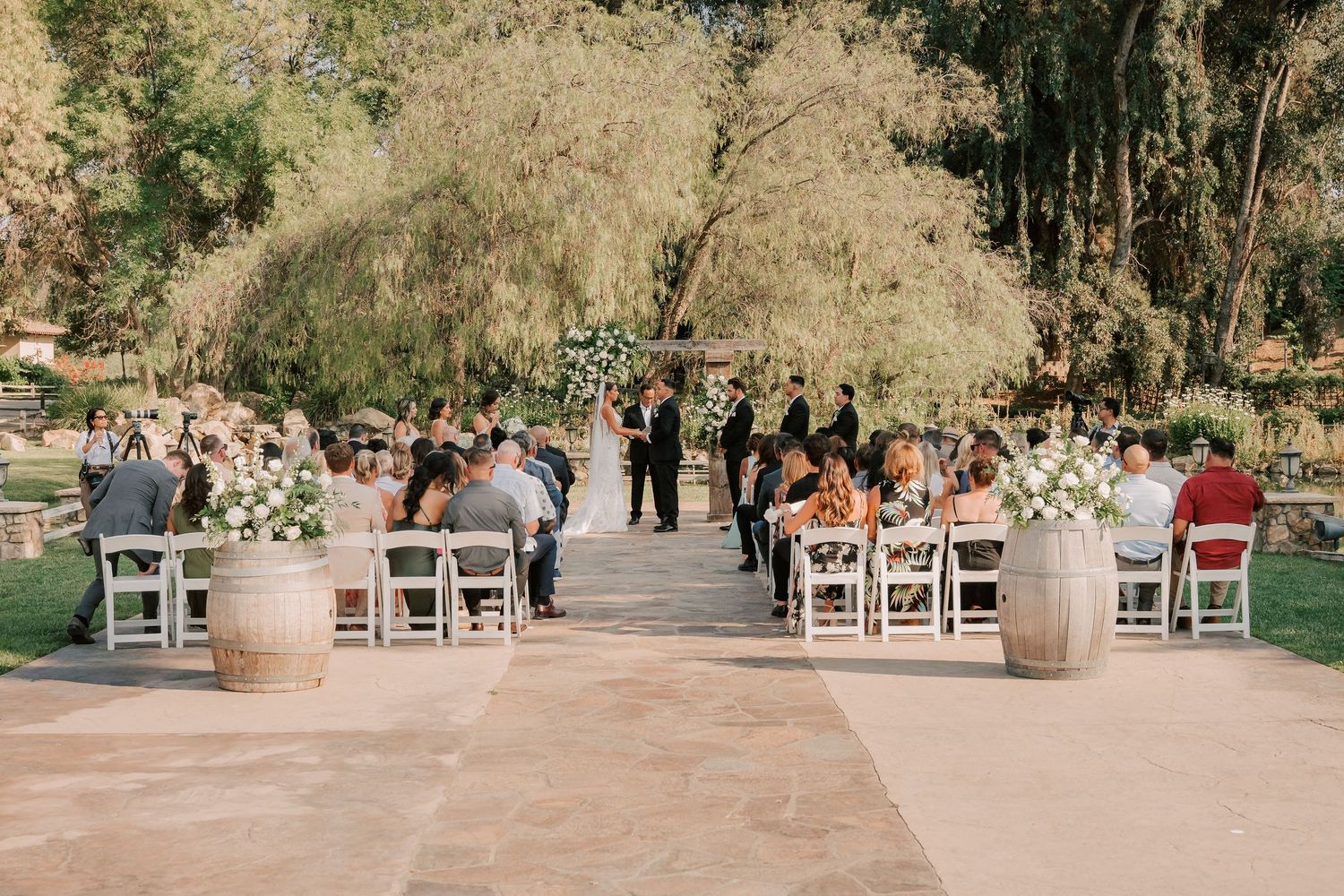 Outdoor wedding ceremony with guests seated in white chairs on a stone patio surrounded by lush green trees and floral arrangements.