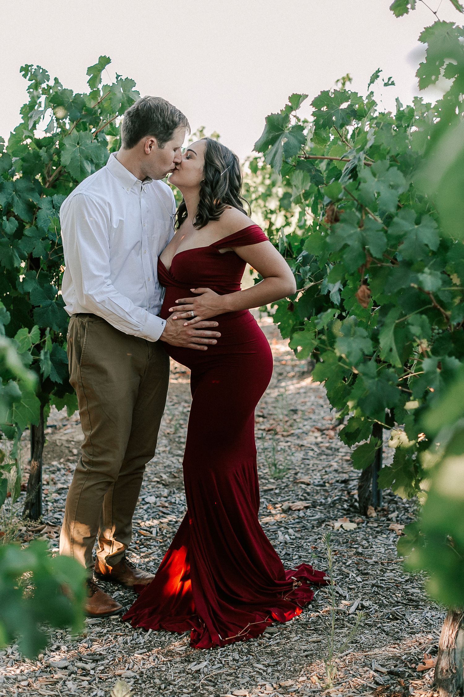 Couple sharing a kiss in a vineyard, woman in elegant red maternity gown, man in white shirt and khakis.