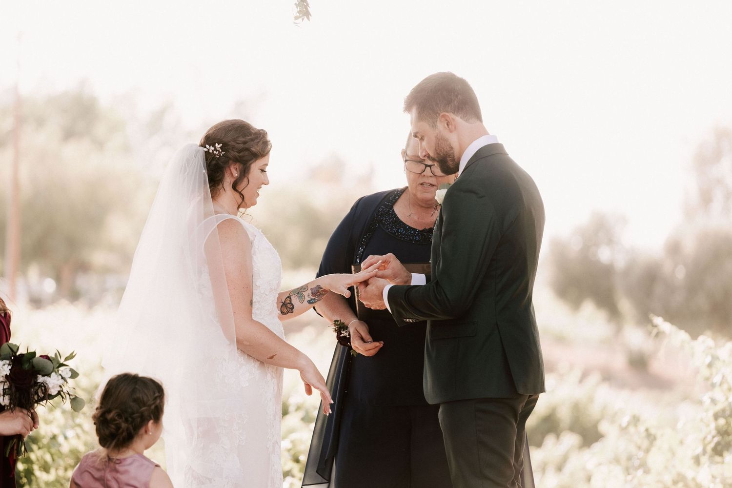 Bride and groom exchanging rings during outdoor wedding ceremony with officiant, soft sunlit background.