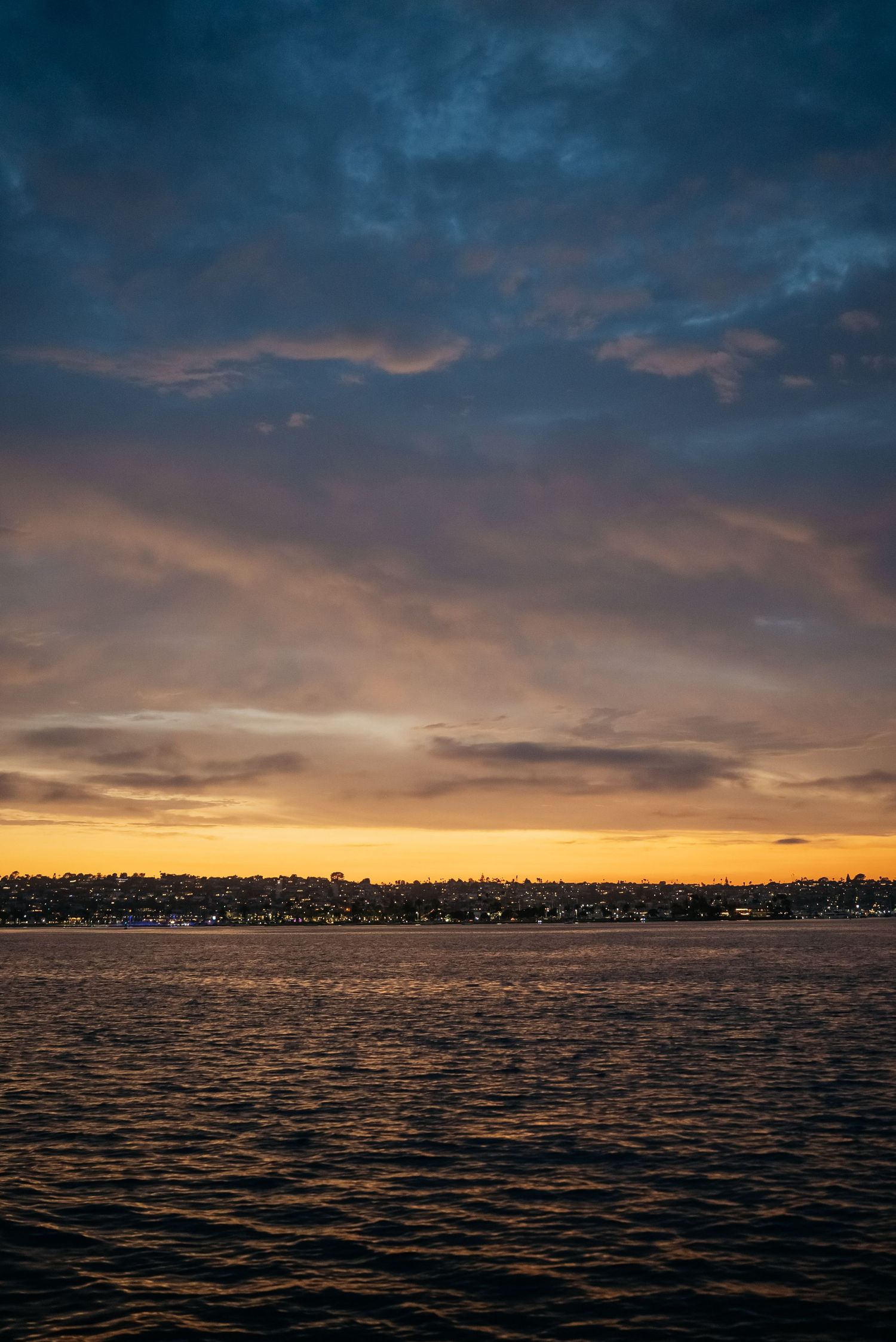 Dramatic golden sunset over ocean with dark blue clouds and glowing horizon over coastal city.