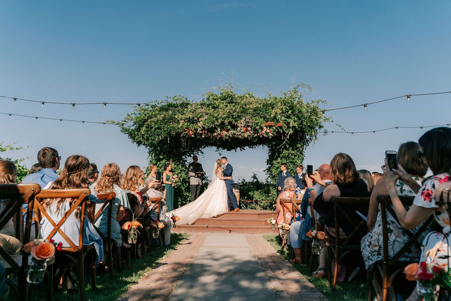 Outdoor wedding ceremony with bride and groom at floral arch altar, guests seated on both sides under string lights on sunny day.