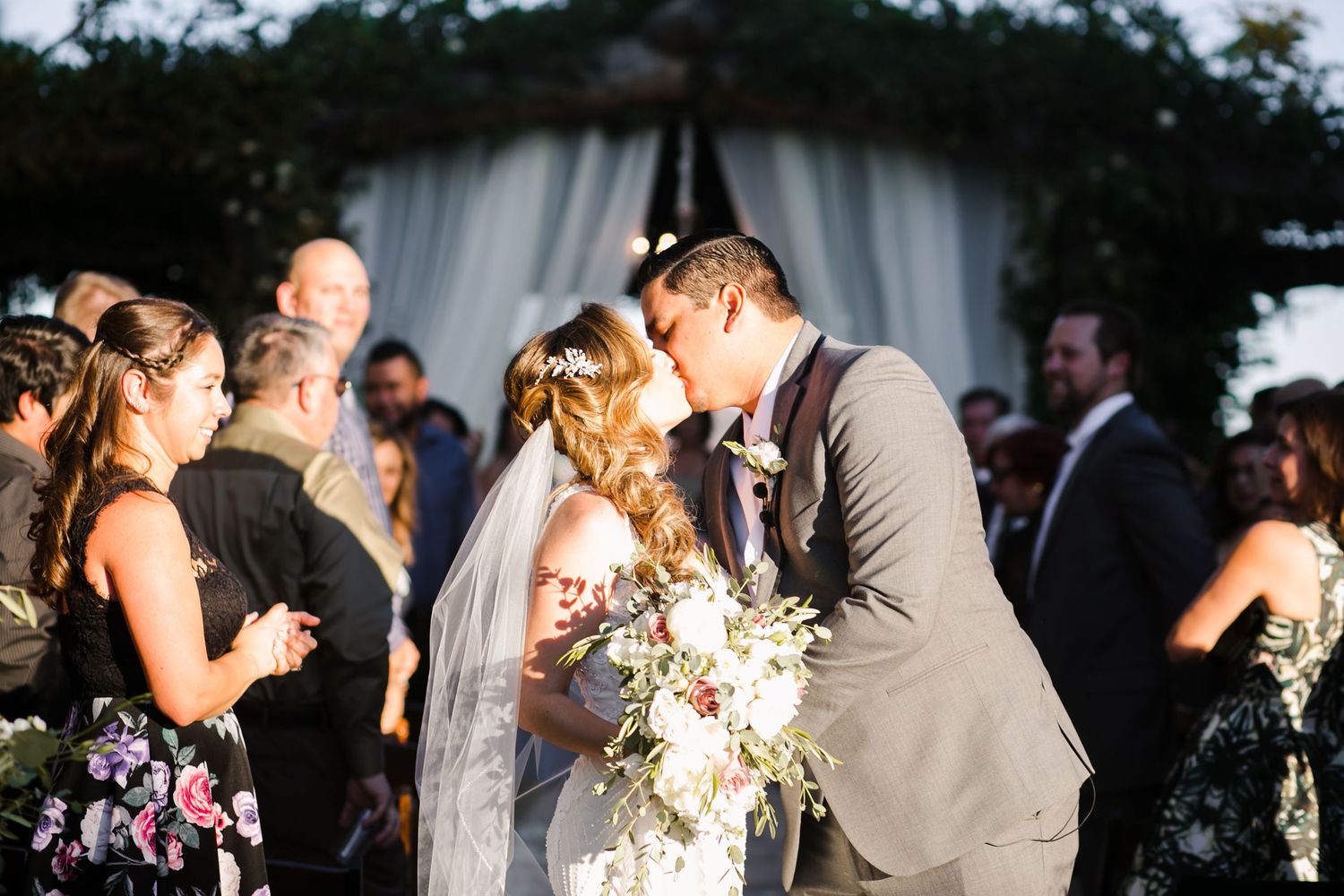 Bride and groom share first kiss at outdoor wedding ceremony with bridesmaids and guests celebrating in background.