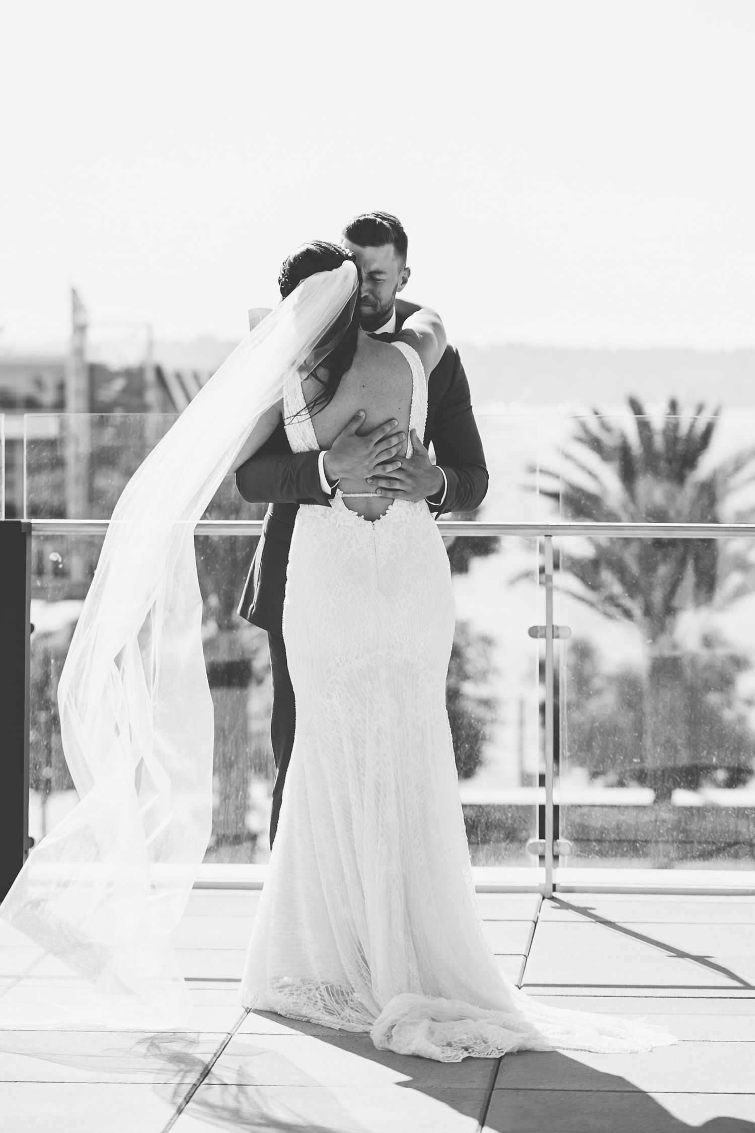 Black and white photo of a bride and groom embracing on a rooftop terrace, with palm trees in the background.