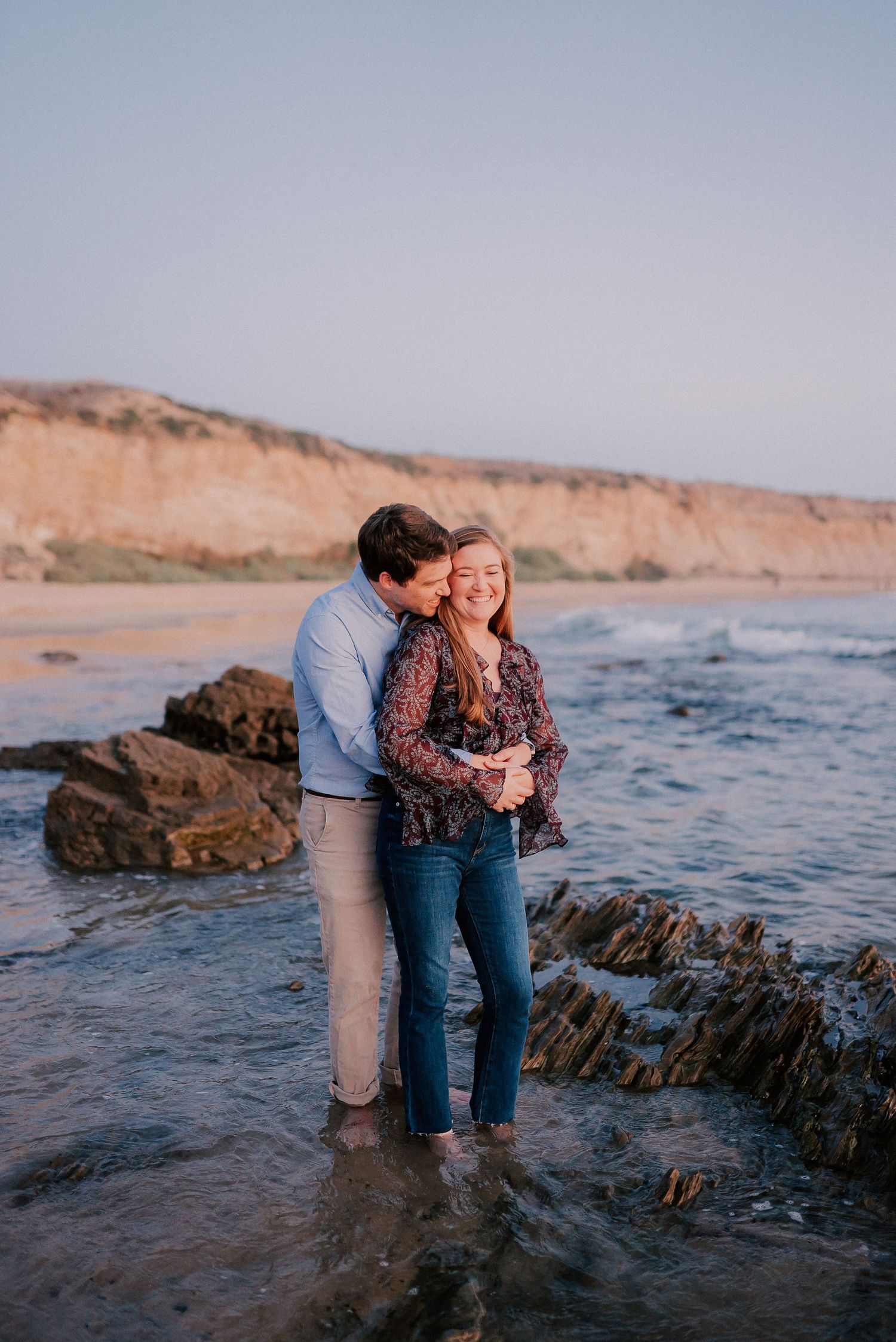 Couple embracing on rocky beach at sunset with golden cliffs in background, standing in shallow water.