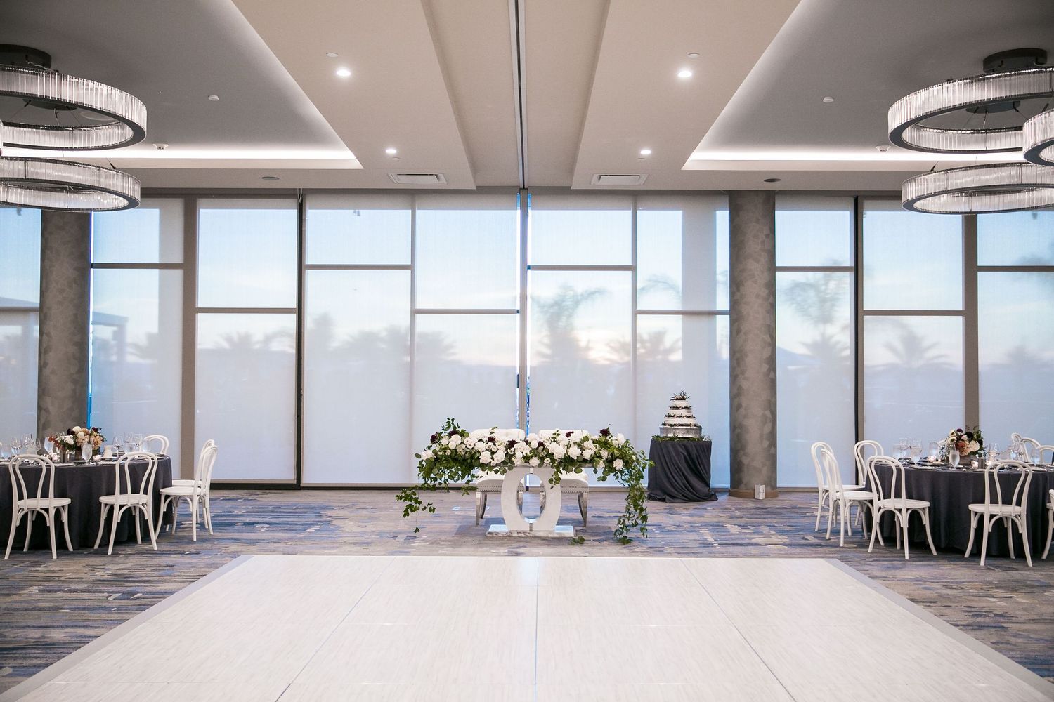 Elegant wedding reception ballroom with white dance floor, floral centerpiece, round tables, and large windows with sheer drapes.