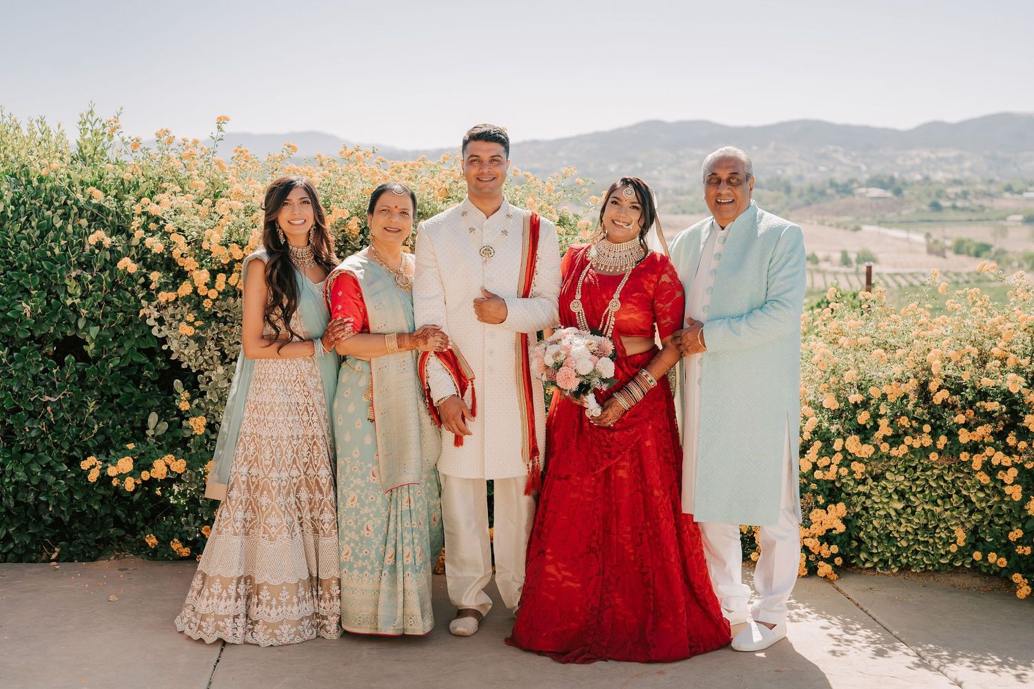 Indian wedding family portrait with bride in red lehenga and groom in white sherwani before yellow roses.