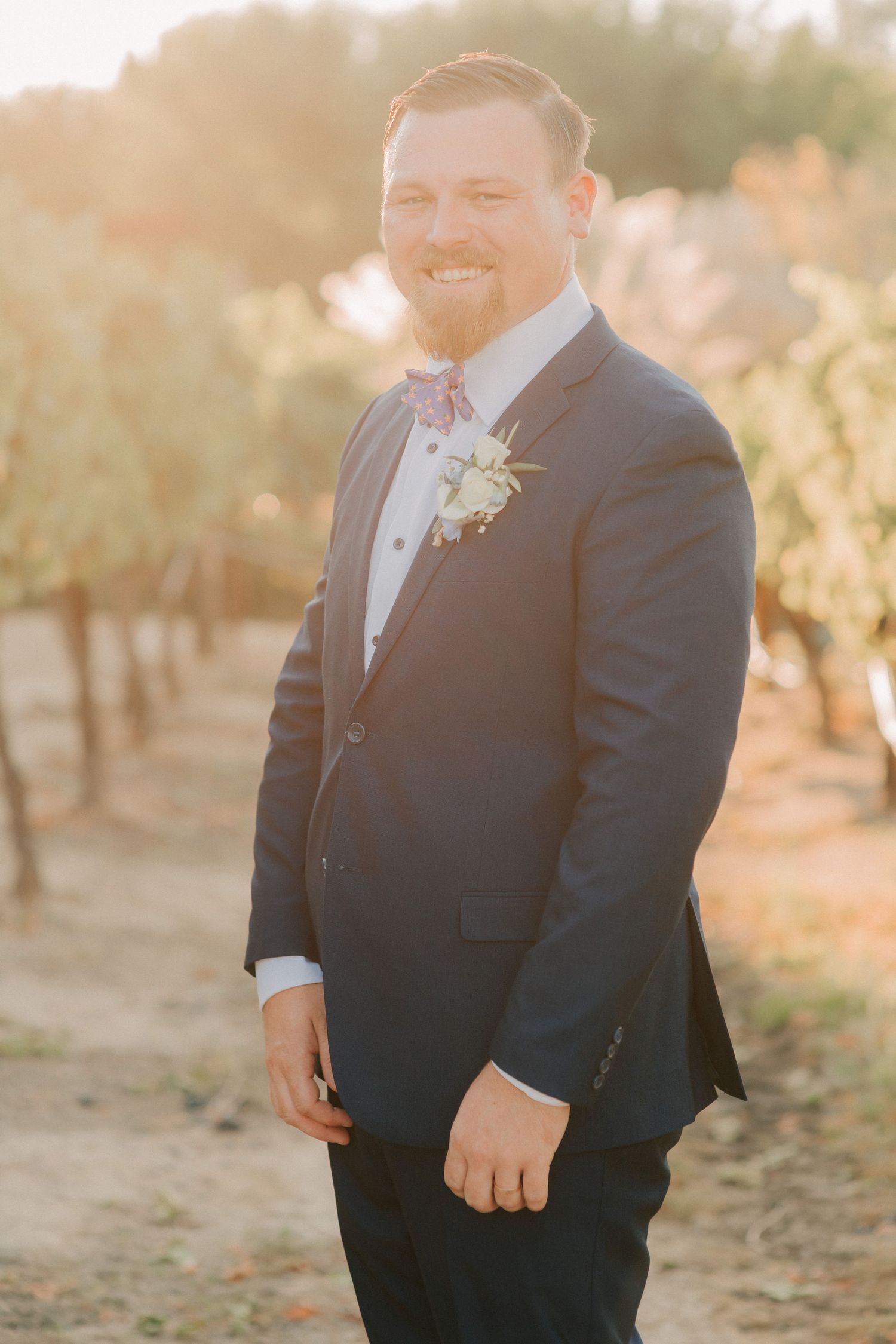 Smiling groom in navy suit with floral bow tie and boutonniere posing outdoors in golden hour sunlight.