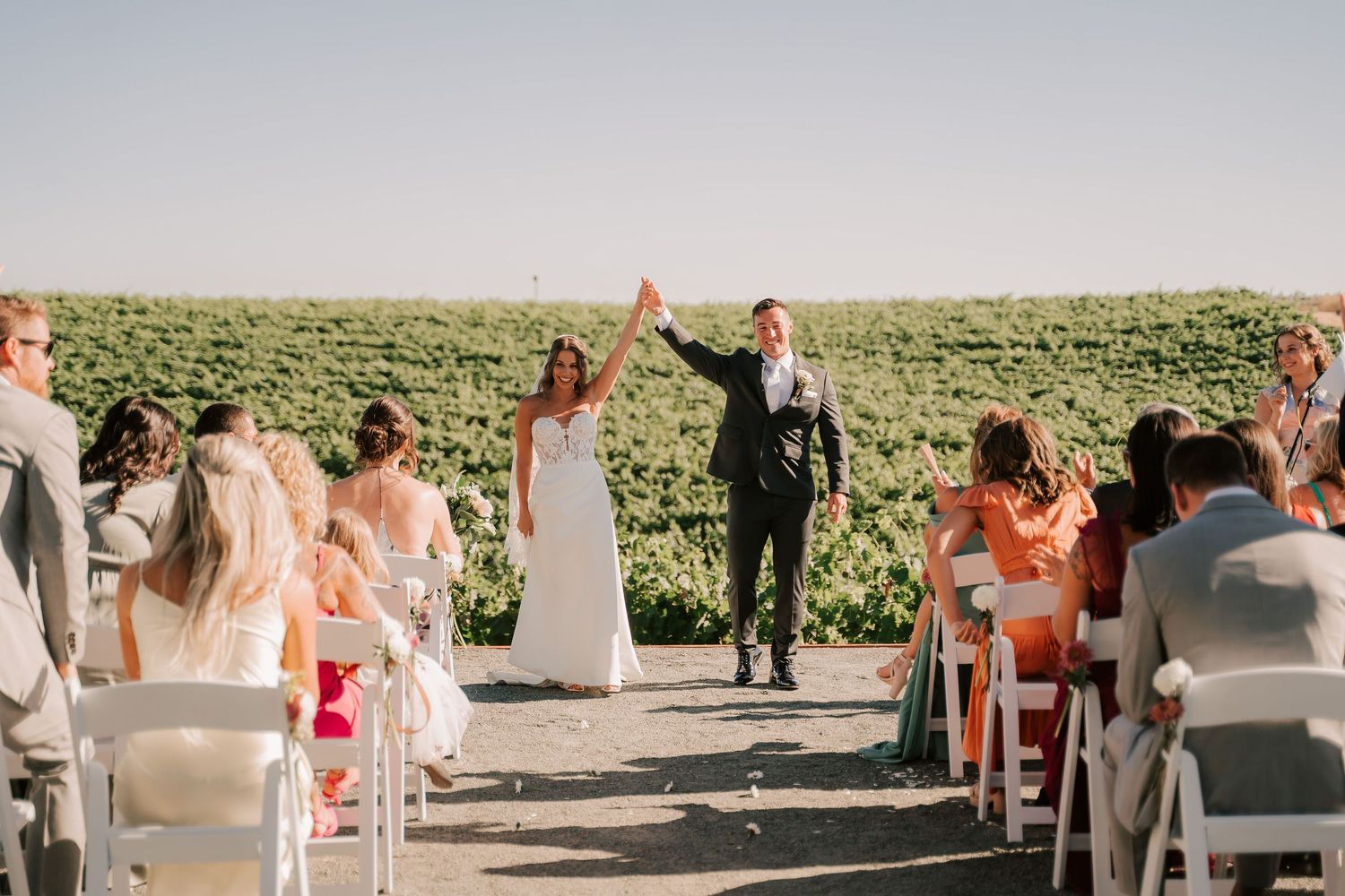 Joyful newlyweds raise hands walking down outdoor wedding aisle lined with guests in white chairs.