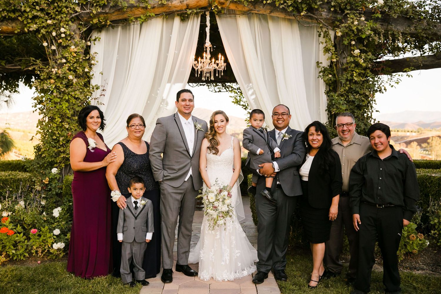 Wedding party poses under floral chandelier arch outdoors, bride in white gown with bouquet, groom in gray suit, family surrounding them.