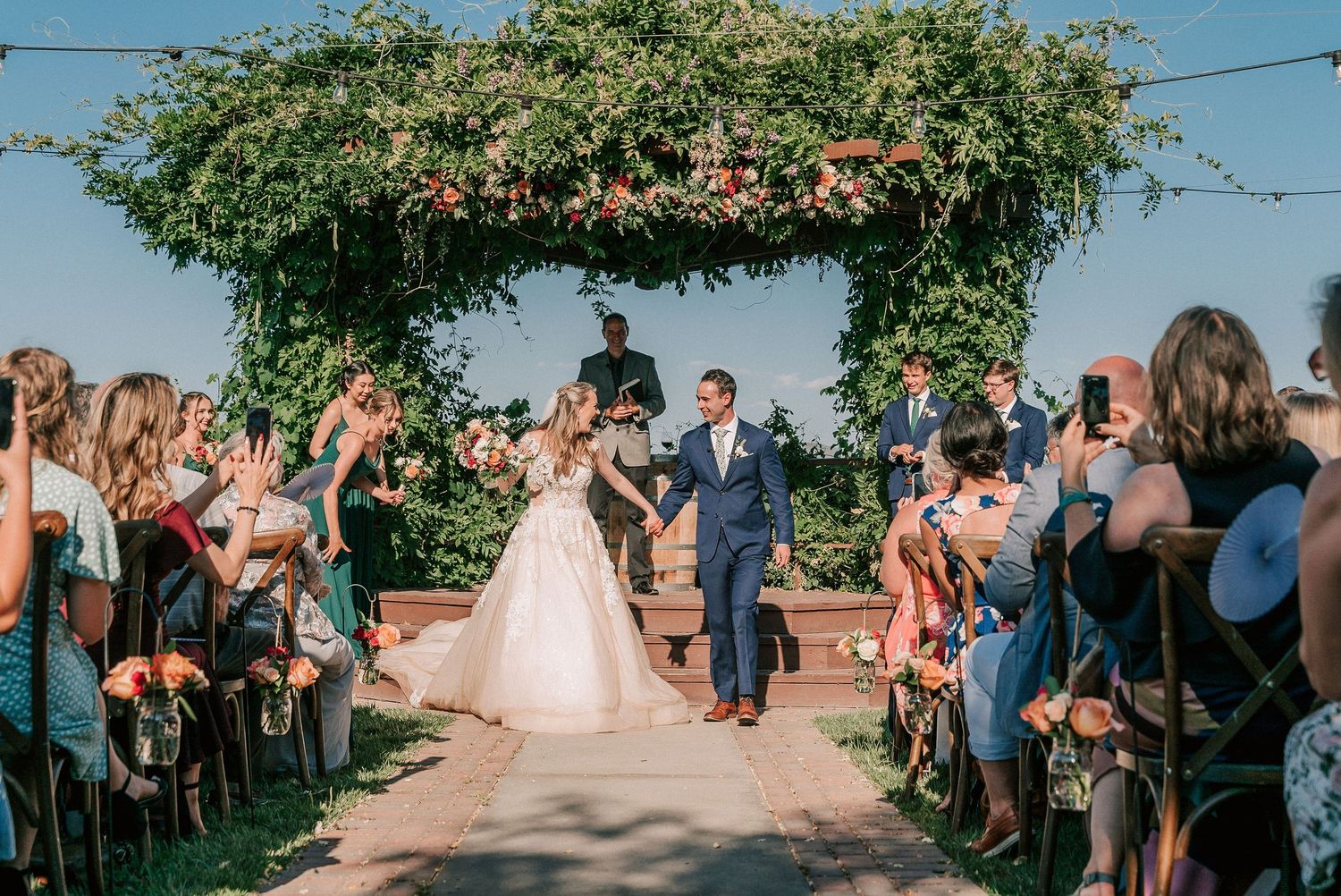 Newlywed couple walks down outdoor wedding aisle under lush green floral arch, guests cheering and photographing the joyful ceremony.