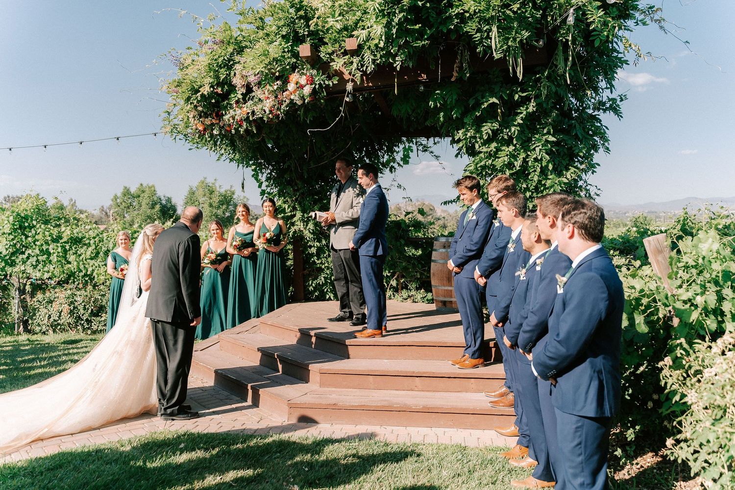 Outdoor vineyard wedding ceremony with bridal party in navy suits and emerald dresses under a floral arch on wooden steps.