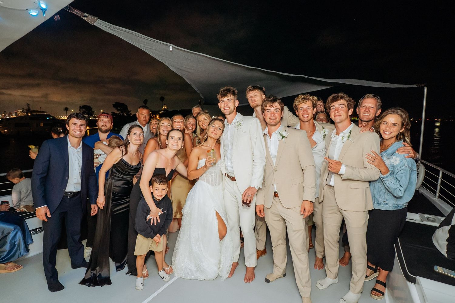 Wedding couple in white attire poses with guests on yacht deck during evening reception celebration.
