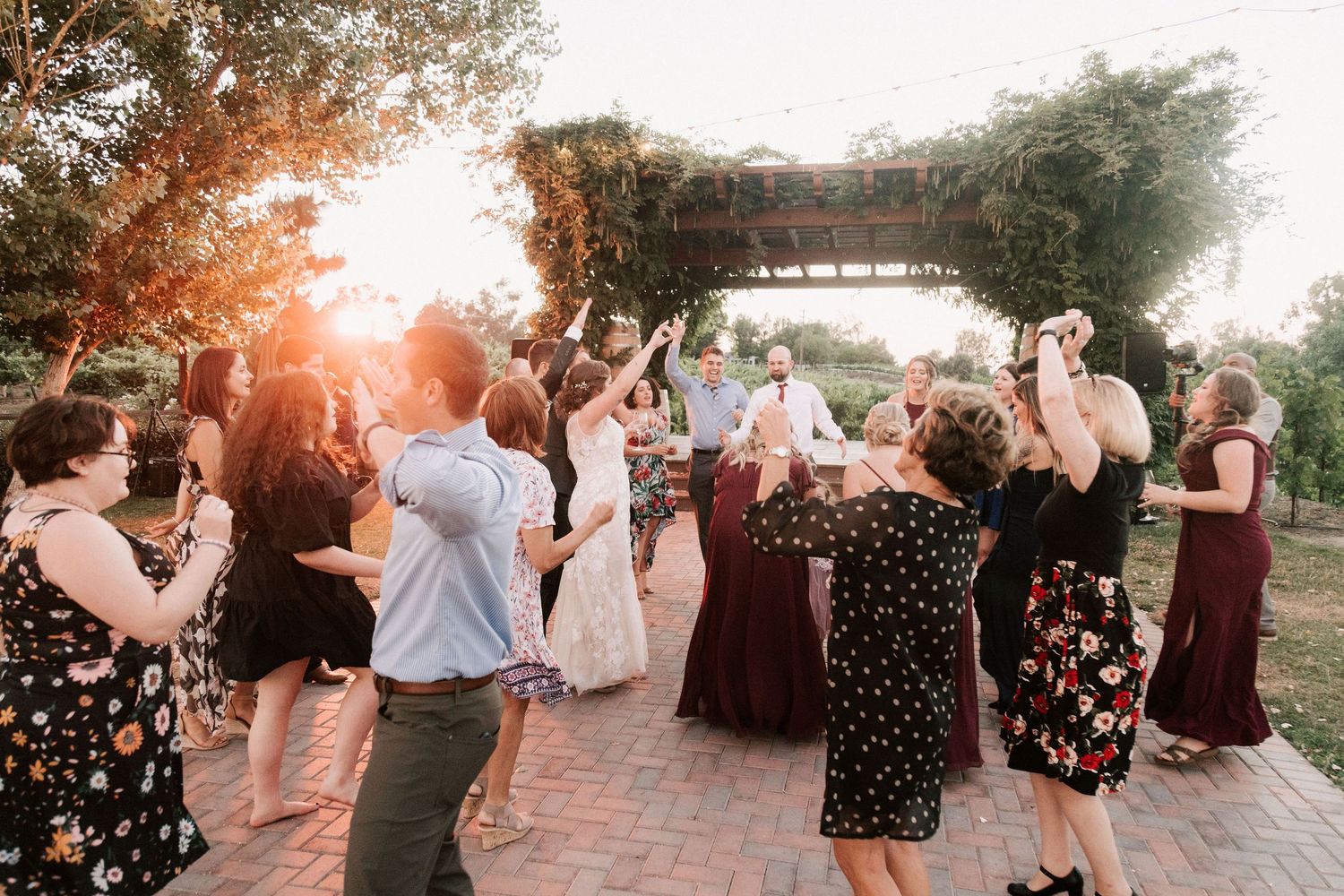 Guests dance and celebrate outdoors at a golden hour wedding reception under a floral pergola.