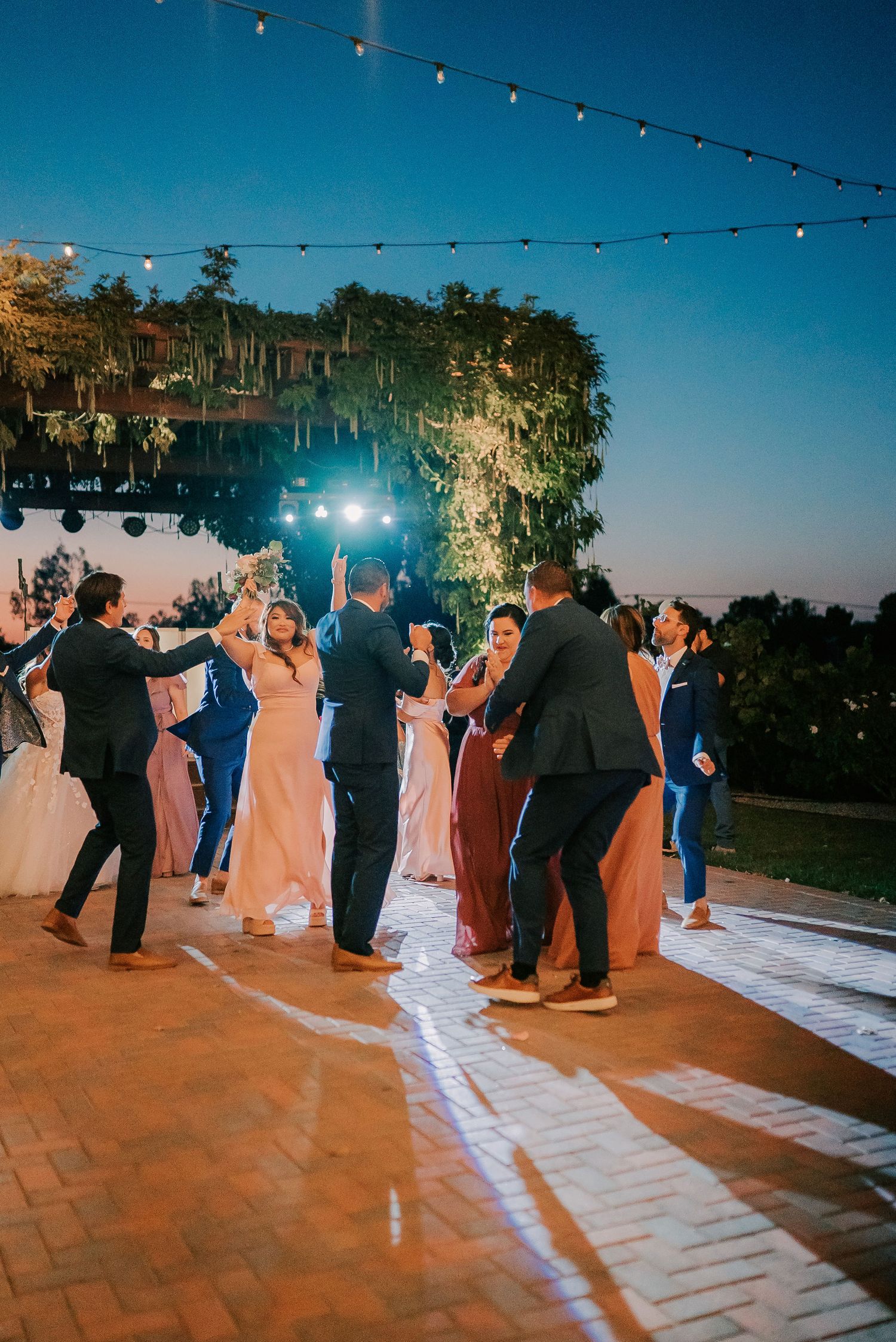 Guests dancing outdoors at a wedding reception under string lights at dusk, with lush greenery in the background.