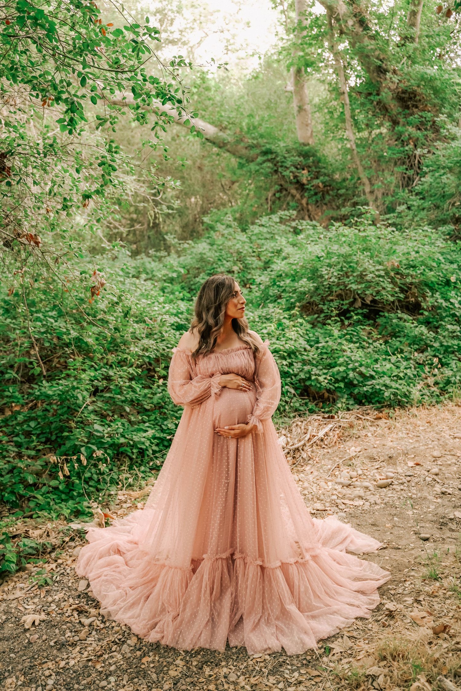 Pregnant woman in flowing pink tulle maternity gown poses outdoors surrounded by lush green foliage.