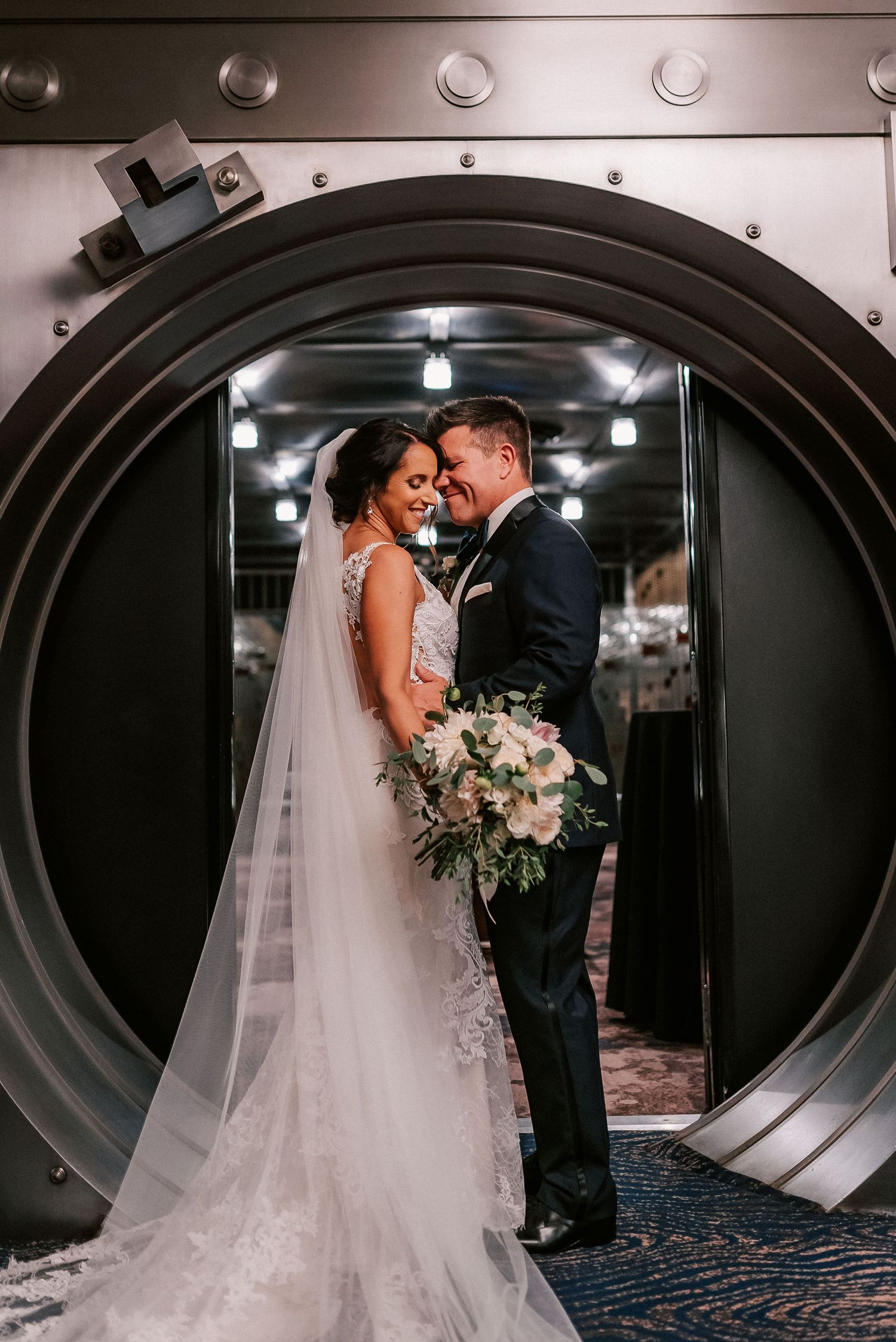 Bride and groom share romantic moment framed by ornate vault door, bride holding white floral bouquet in elegant lace gown.
