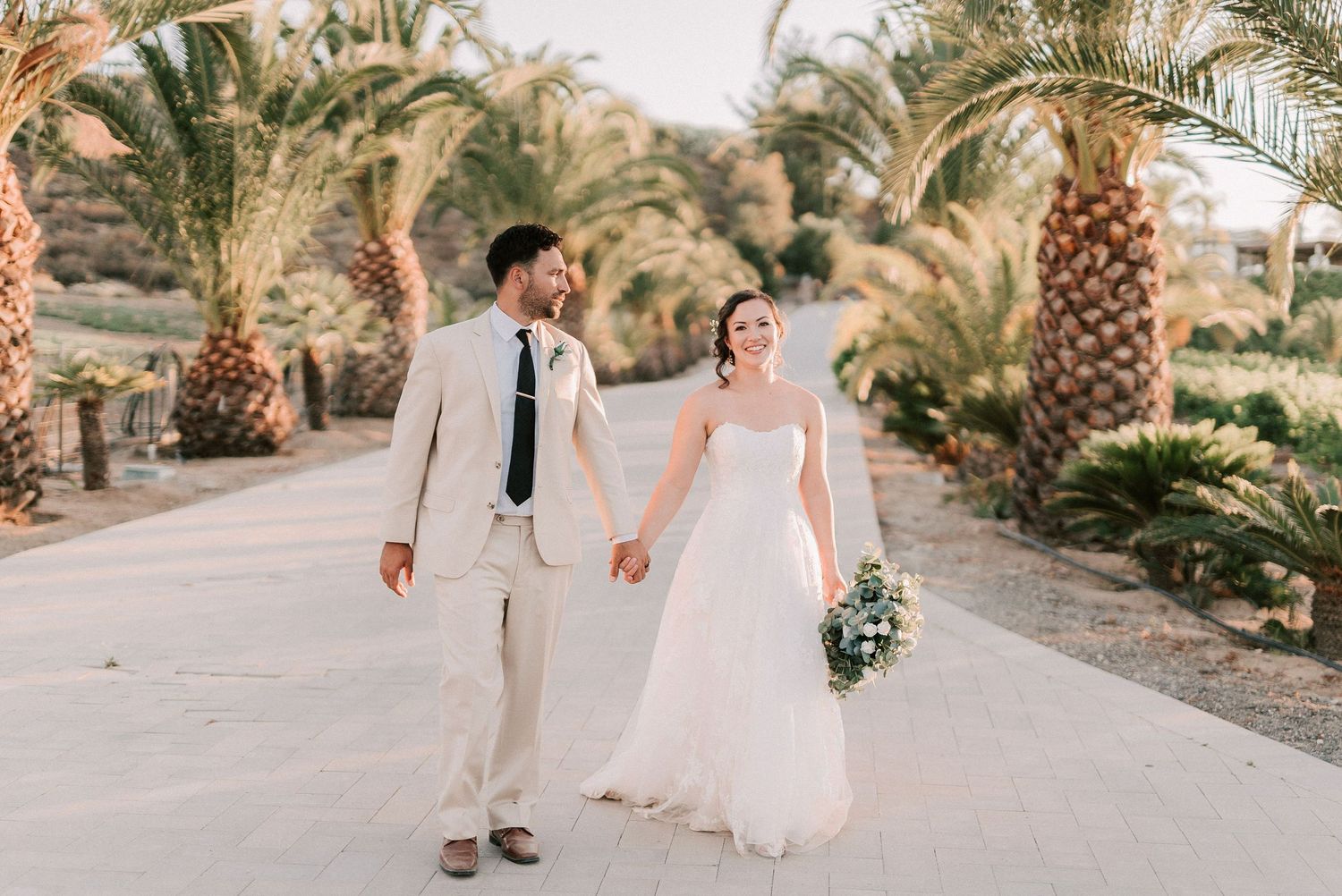 Bride and groom walk hand-in-hand along a sunny palm-lined path, bride in white gown holding blue bouquet.