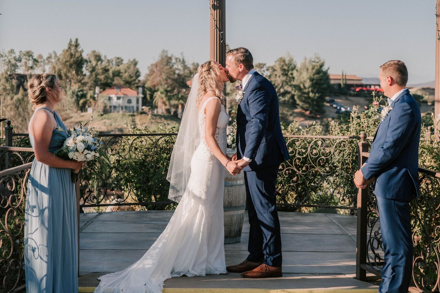 Bride and groom share first kiss at outdoor wedding ceremony on elegant balcony with scenic views.