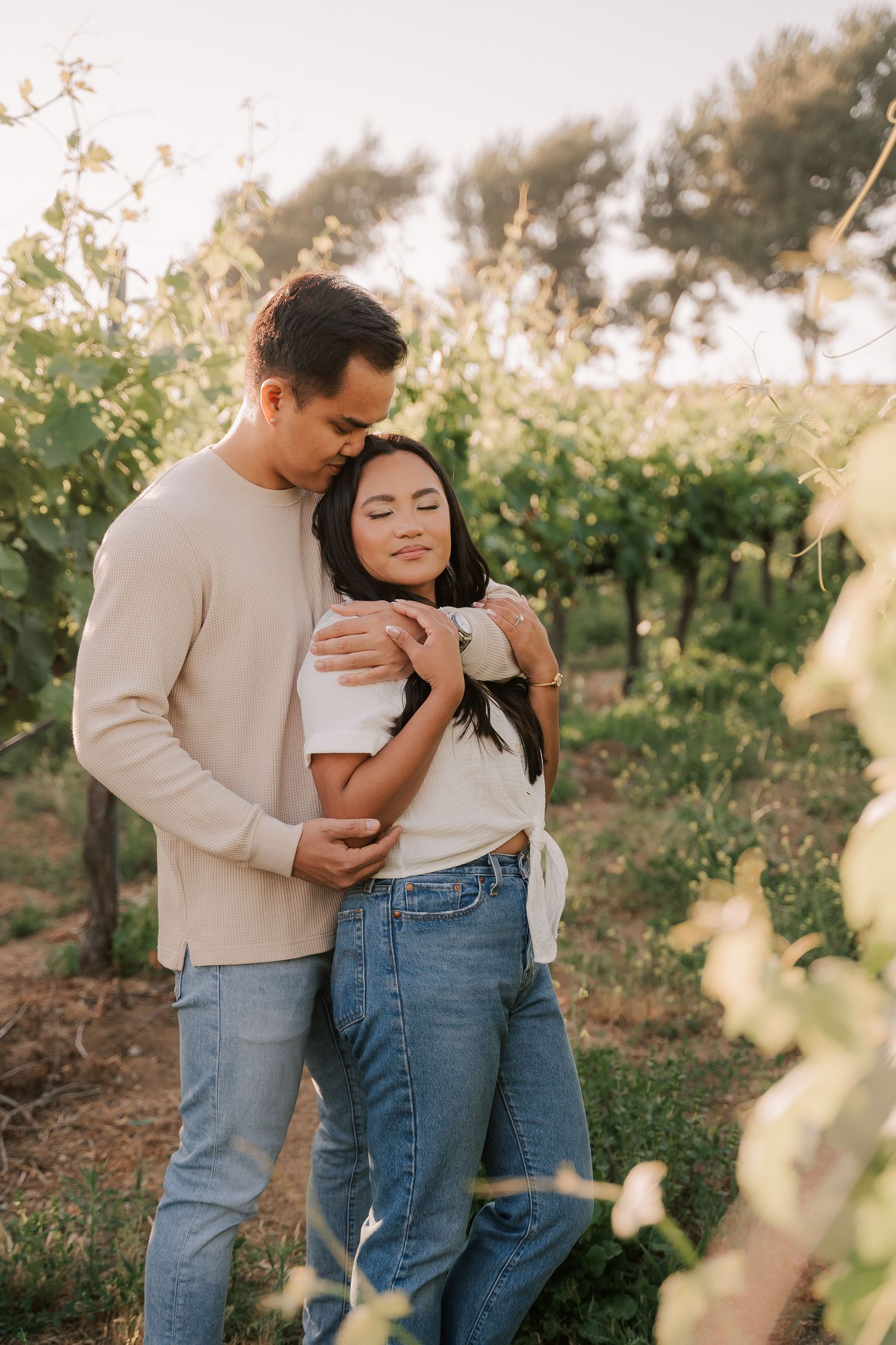 Couple embracing in a sunlit vineyard, wearing casual jeans and neutral tops, surrounded by lush green grapevines.