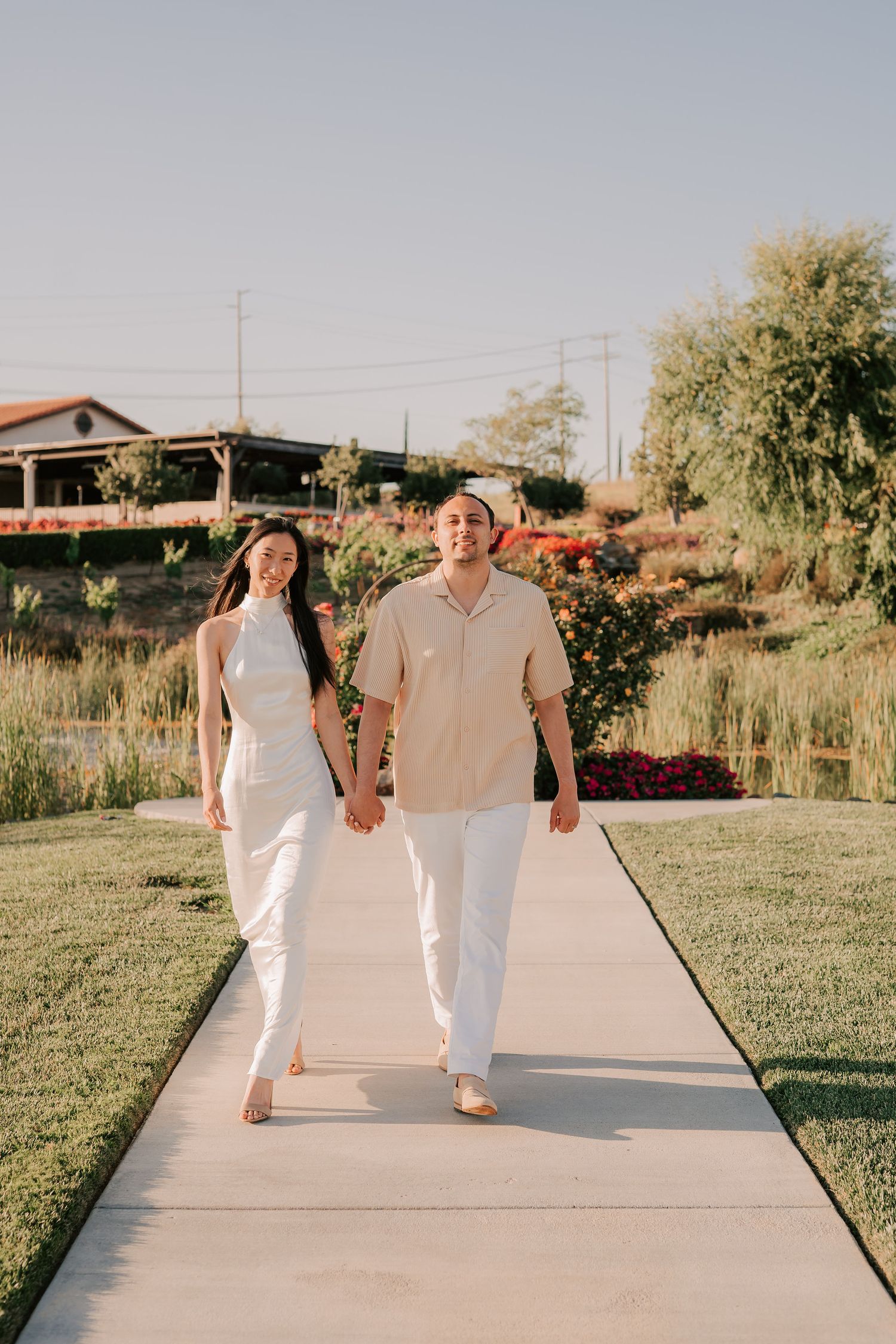 Couple in white outfits holding hands walking on a garden pathway with lush greenery and red flowers.