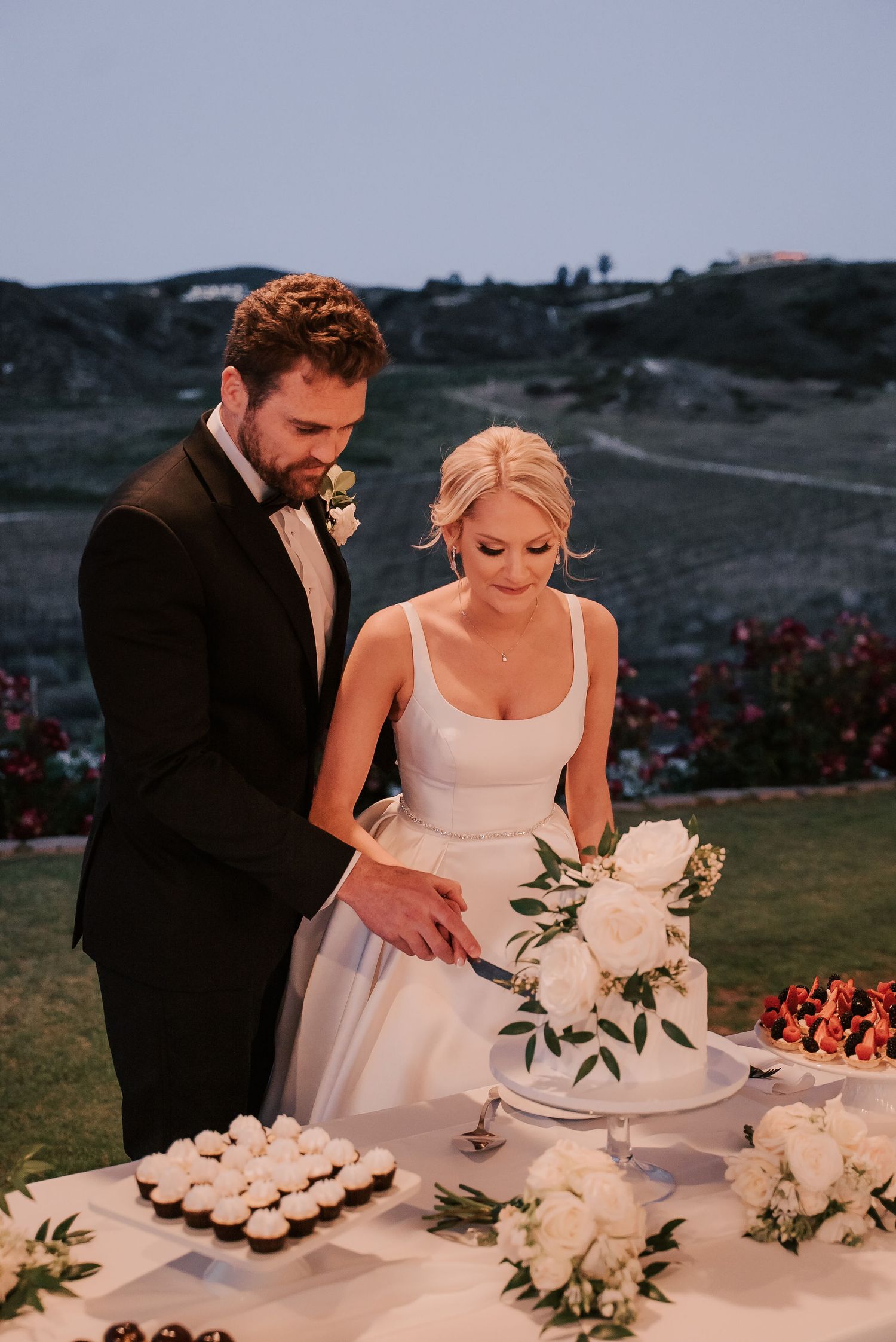 Bride and groom cutting white floral wedding cake outdoors at dusk, with cupcakes and roses on the table.