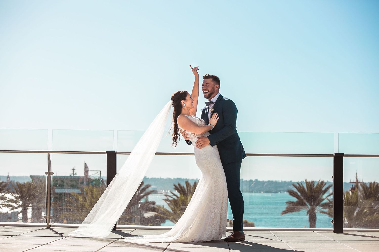 Bride and groom dancing on rooftop terrace with ocean and palm trees backdrop on sunny wedding day.