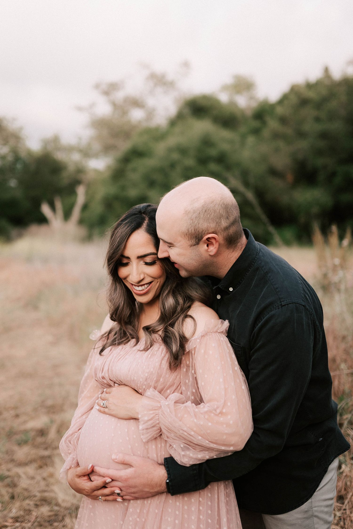 Smiling pregnant woman in pink dress embraced by man in black shirt during outdoor maternity shoot.