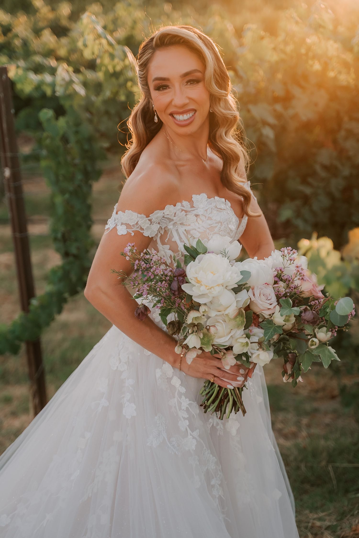 Smiling bride in off-shoulder lace gown holds white and mauve bouquet in sun-kissed vineyard setting.
