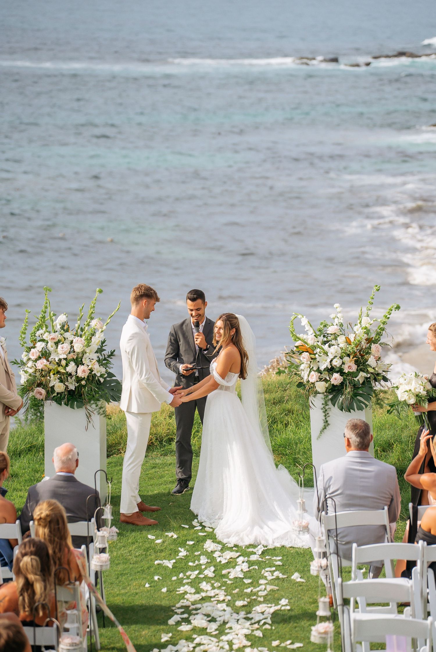 Outdoor oceanside wedding ceremony with bride and groom exchanging vows at floral altar on green cliffside.