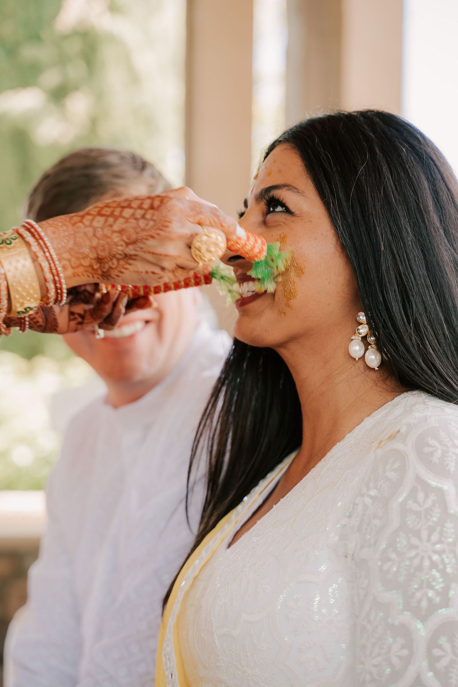 Indian bride with mehndi hands feeds food to woman in white outfit during traditional wedding ceremony ritual.