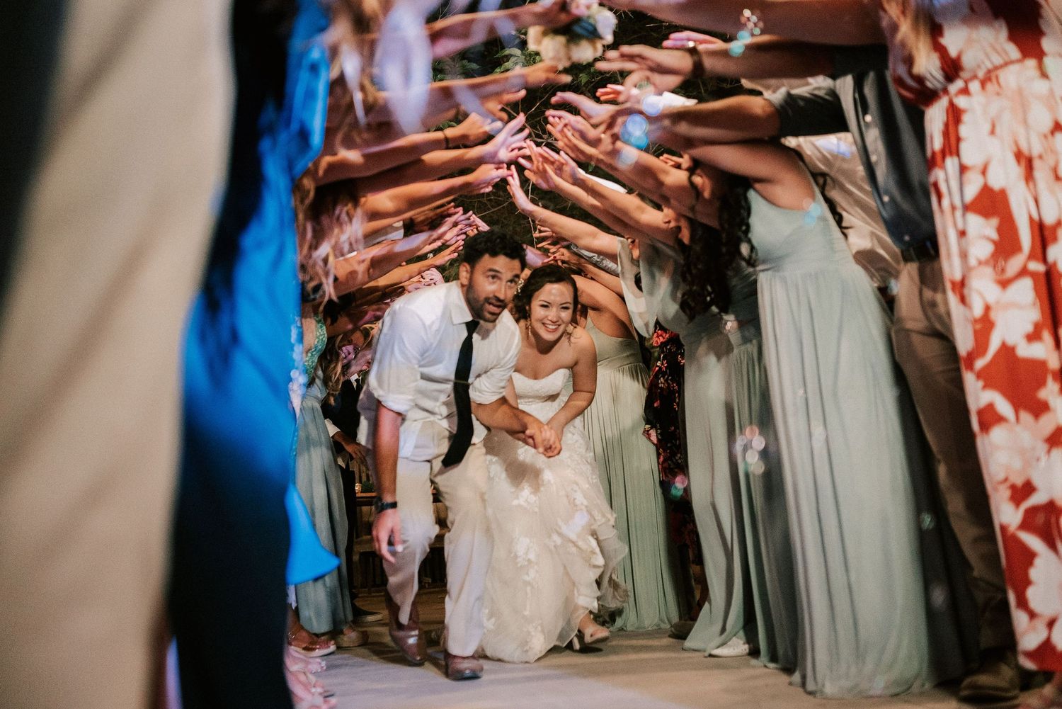 Bride and groom walk through a joyful tunnel of guests tossing flower petals at an outdoor wedding reception.