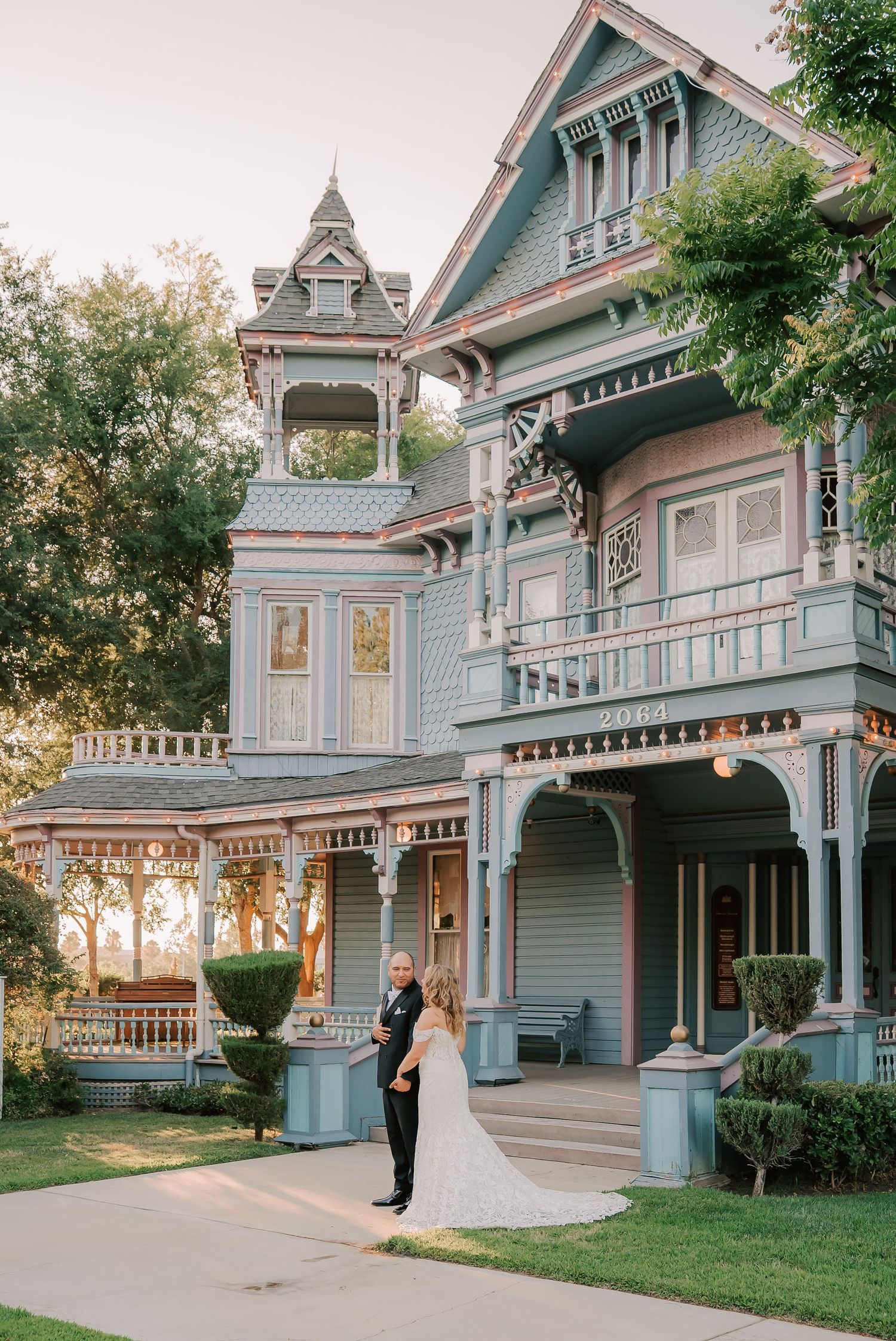 Bride and groom pose before a stunning blue Victorian mansion with ornate details at golden hour.