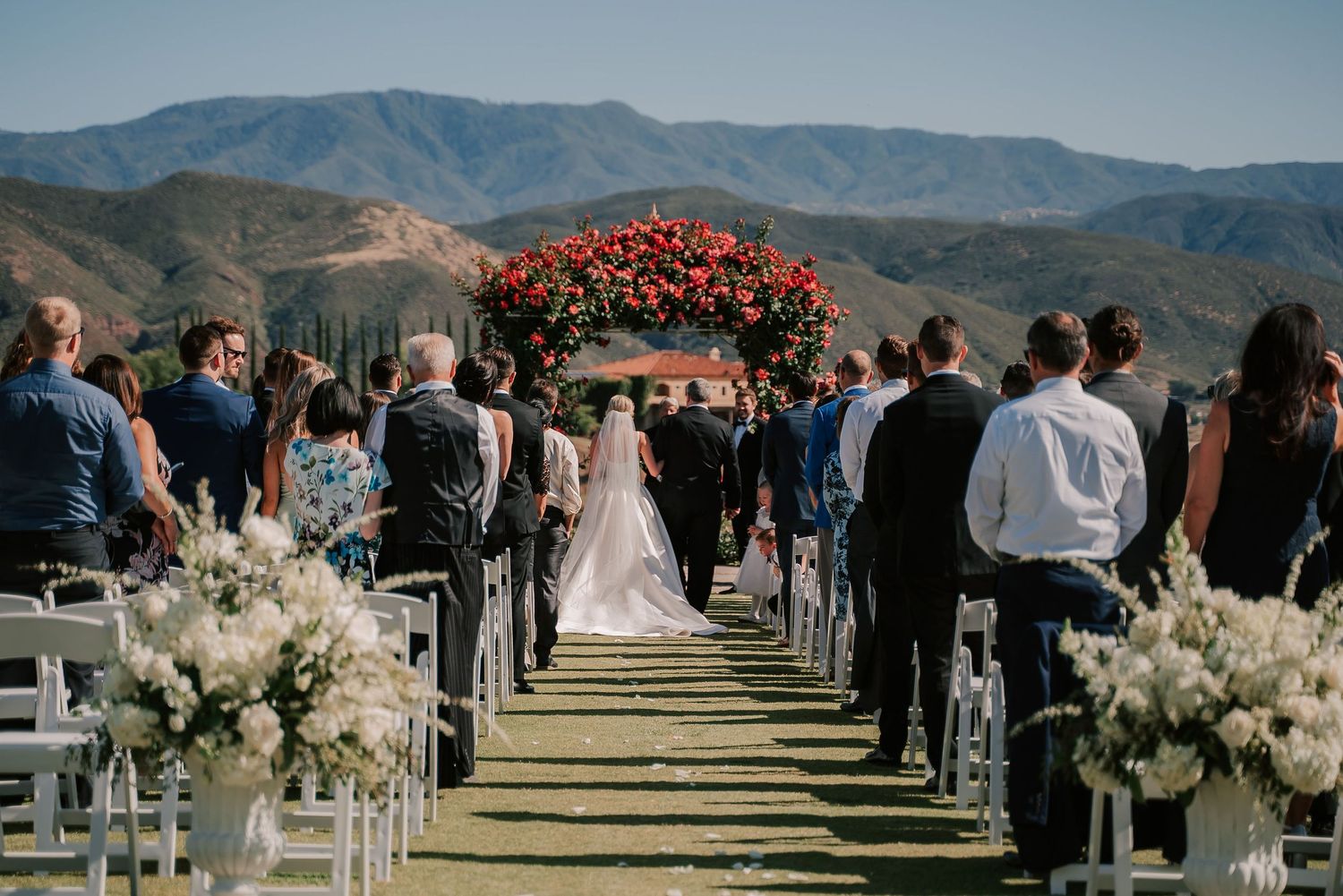 Outdoor wedding ceremony aisle with floral arch, mountain backdrop, and guests seated on white chairs.