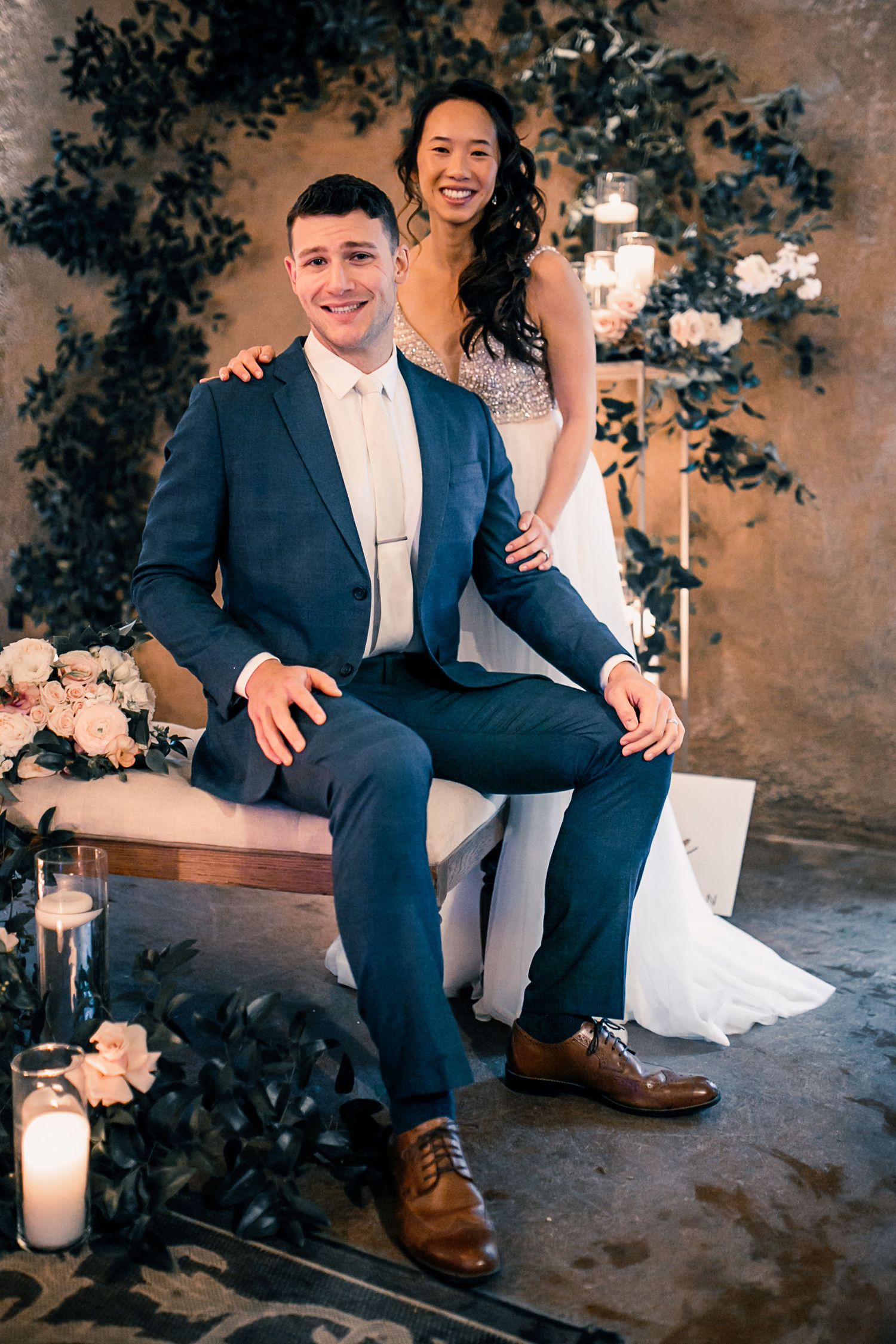 Smiling couple in wedding attire pose together, groom in navy suit seated, bride in white gown standing behind him.