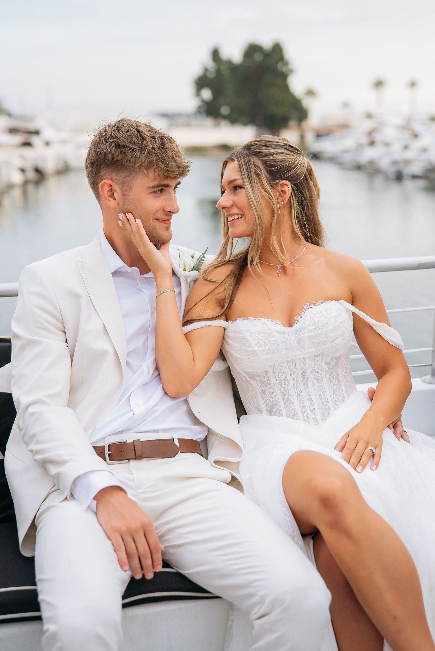 Bride and groom sitting together on a boat at a marina, smiling at each other, wearing white wedding attire.