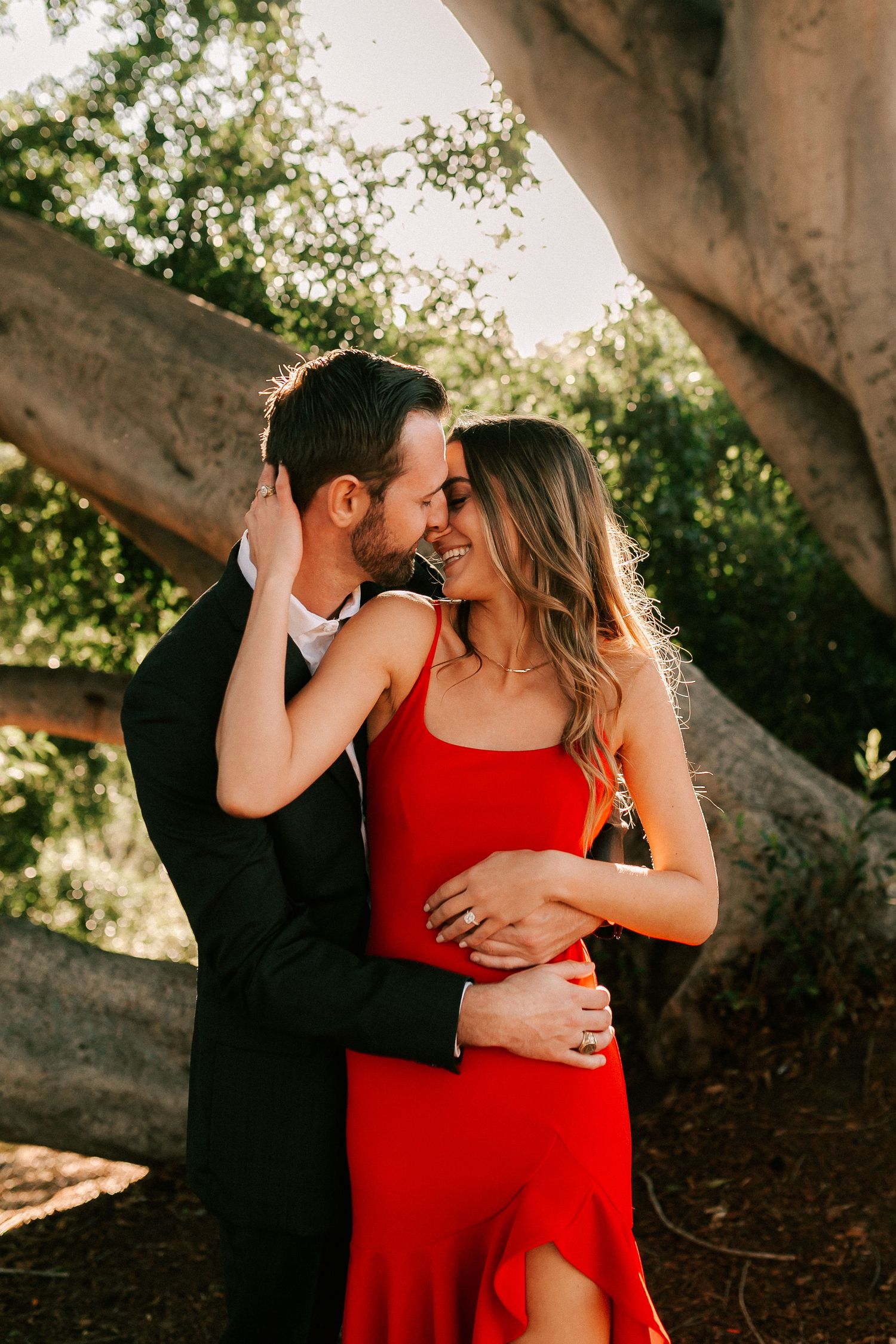 Man in black suit embracing woman in red dress, sharing a tender moment under a large tree outdoors.