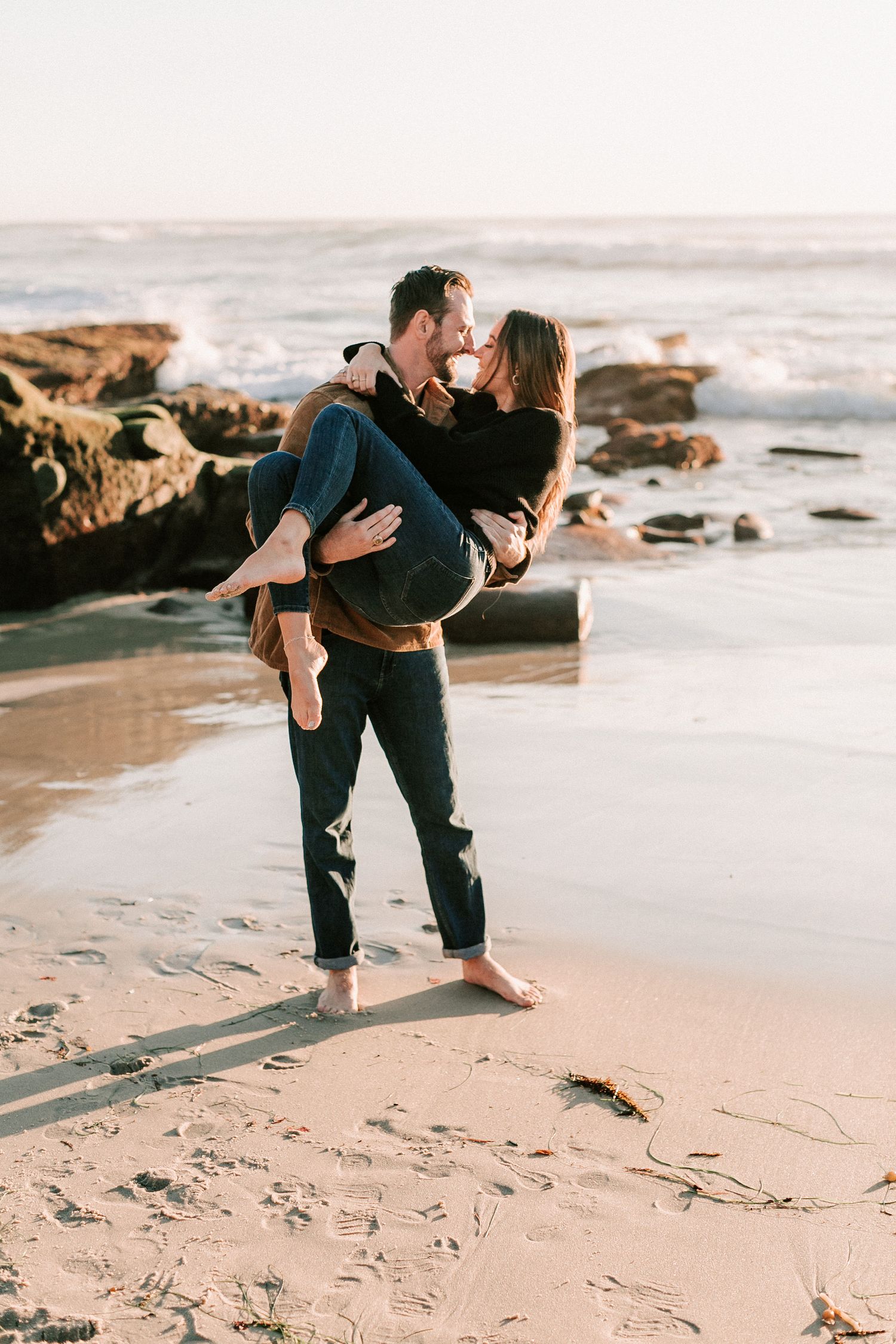 Man carrying woman in arms on rocky beach at golden hour, leaning in for a kiss.