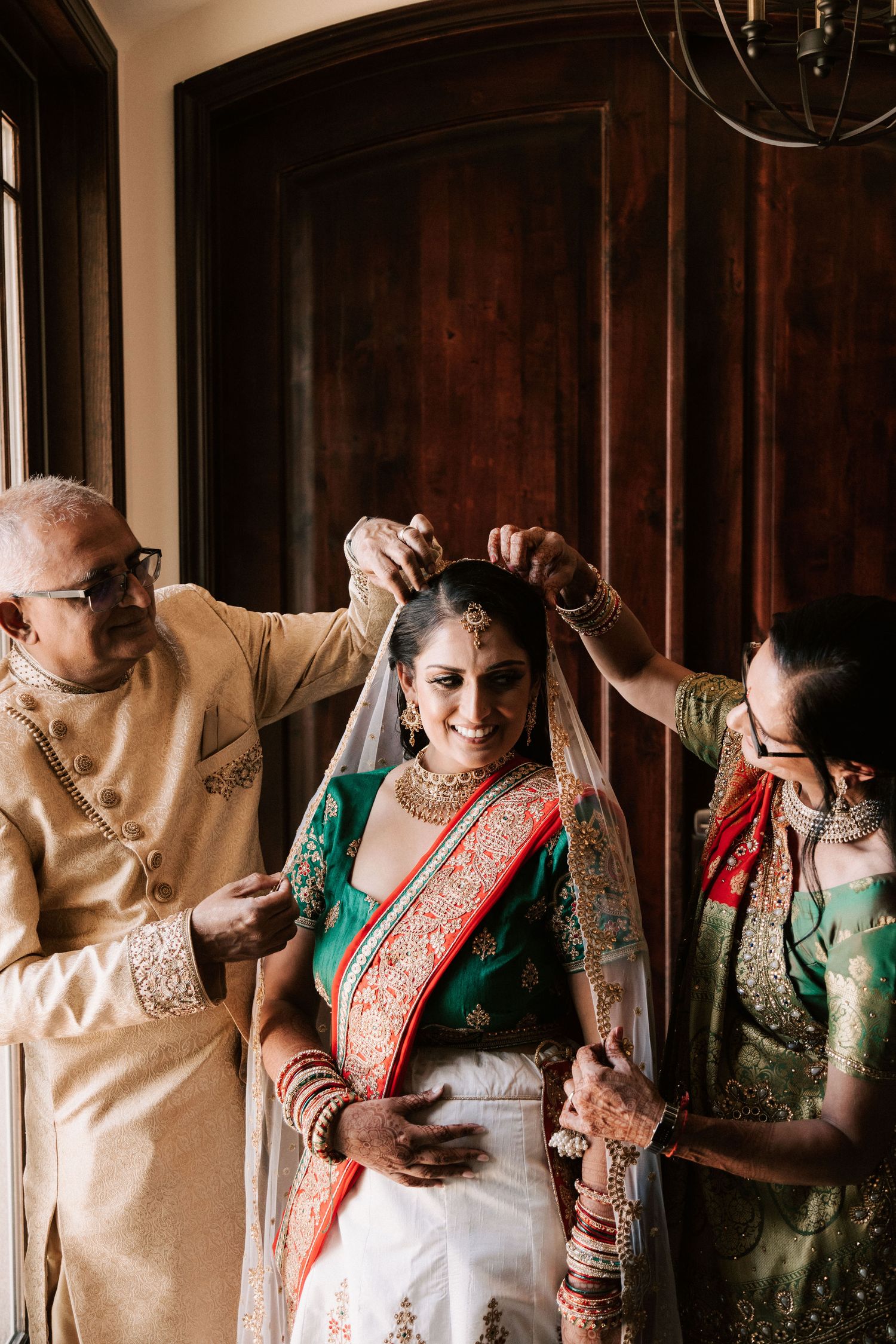 Indian bride smiling as parents place veil during traditional wedding preparation.