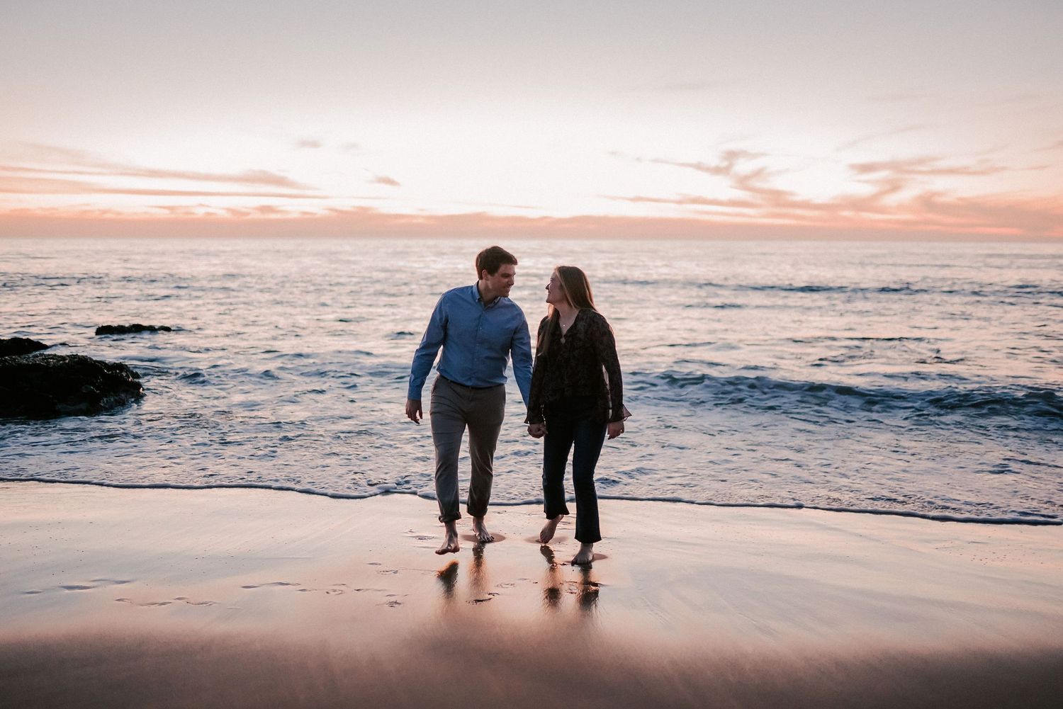 Romantic couple walking hand in hand on sandy beach at colorful sunset with ocean waves.