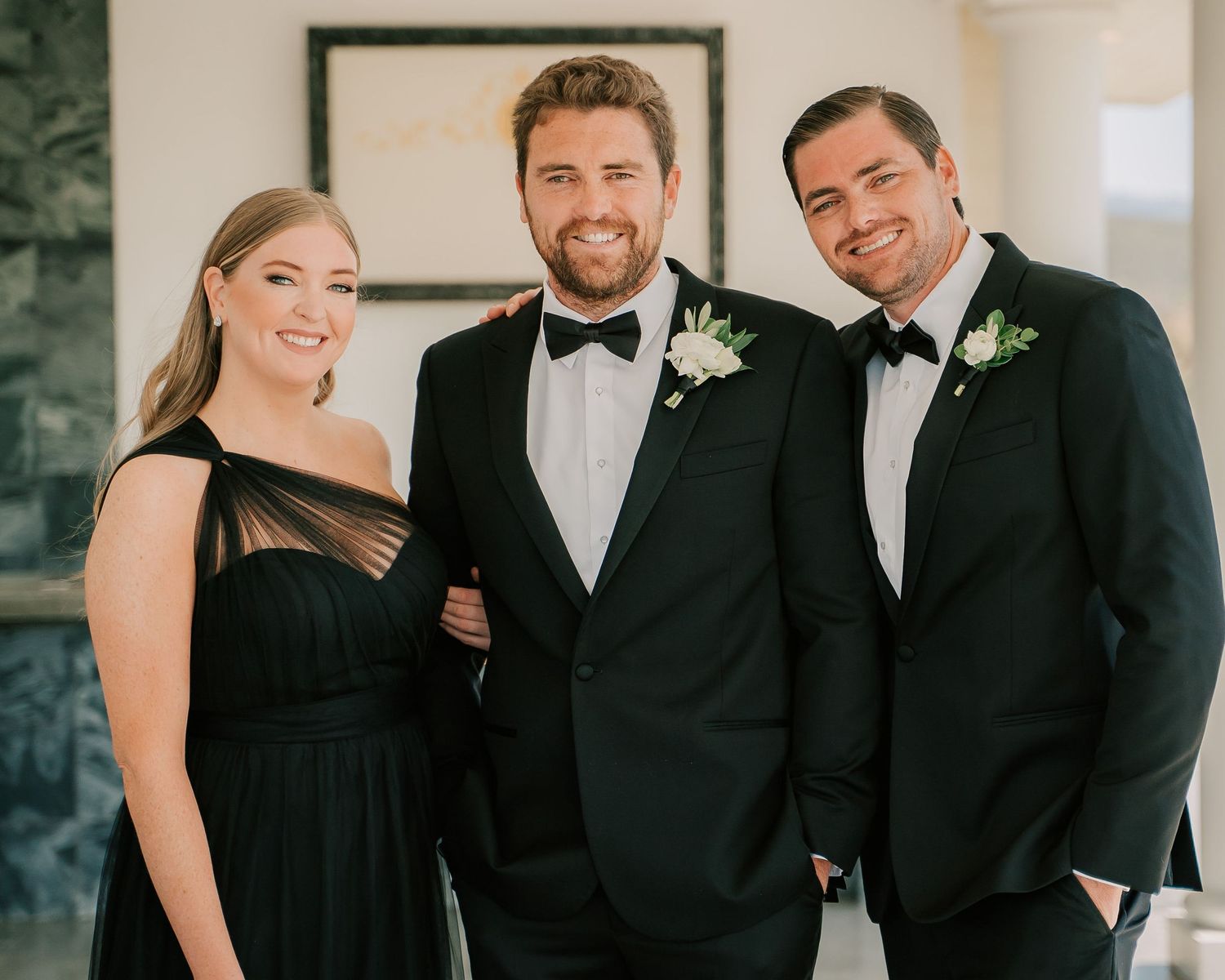 Three elegantly dressed wedding guests in black formal attire smile together, with the groom wearing a boutonniere.
