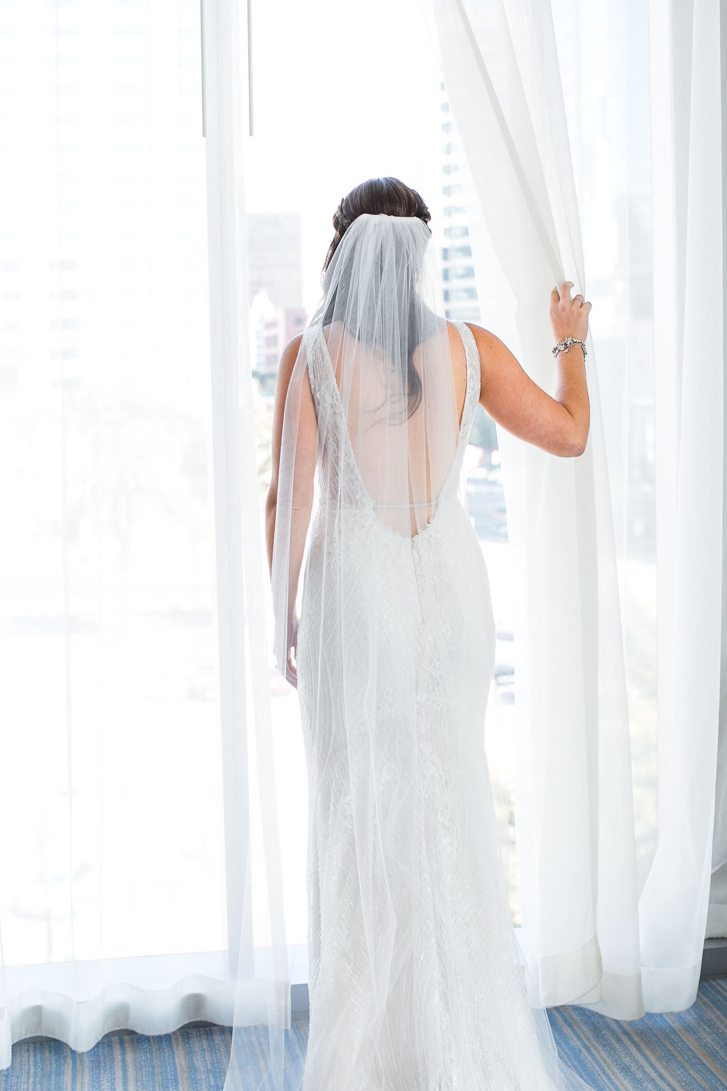 Bride in elegant lace wedding gown and veil stands at bright window, viewed from behind.