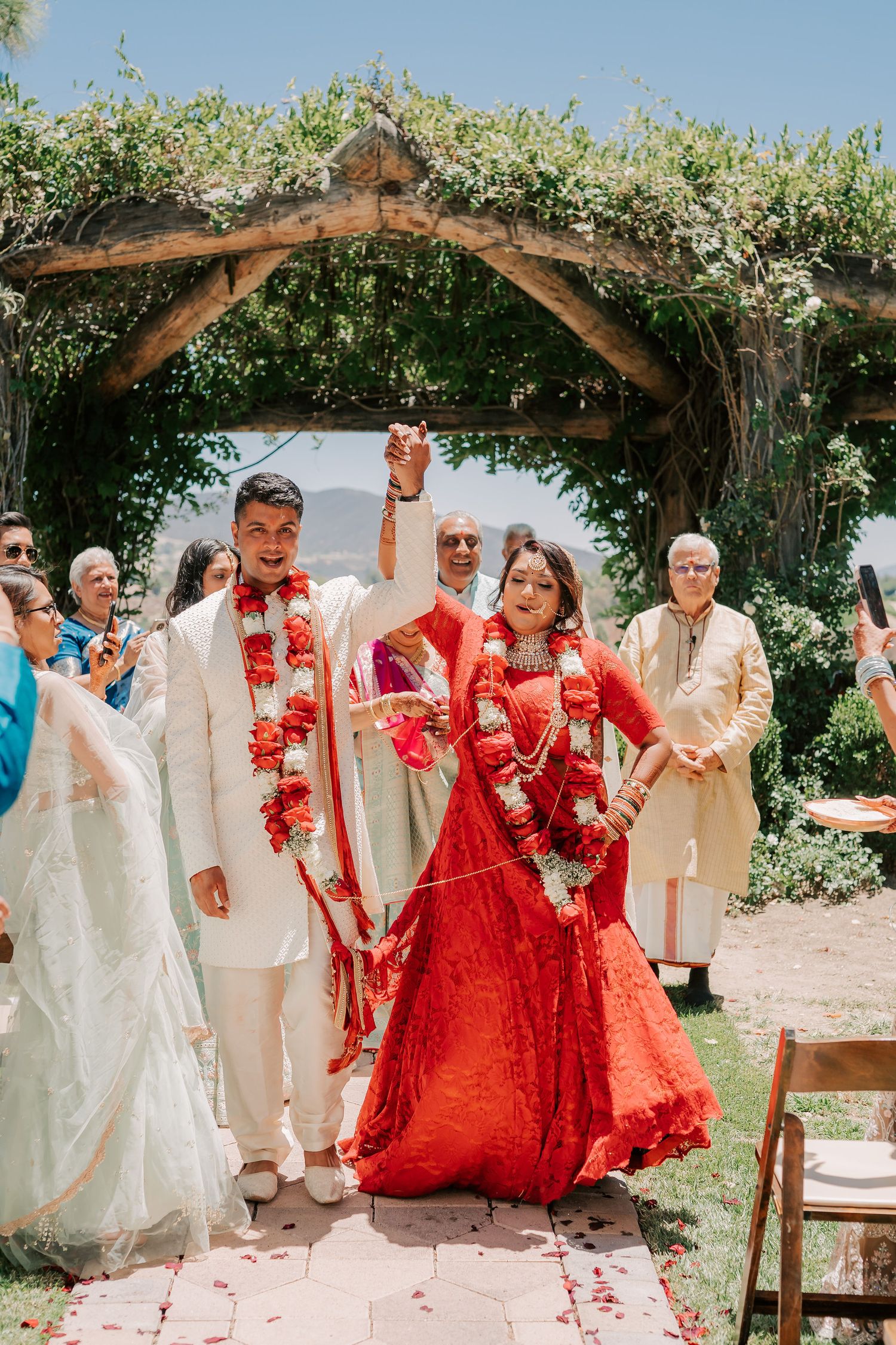 Newlyweds exit ceremony holding hands raised under floral arch in red and white wedding attire.