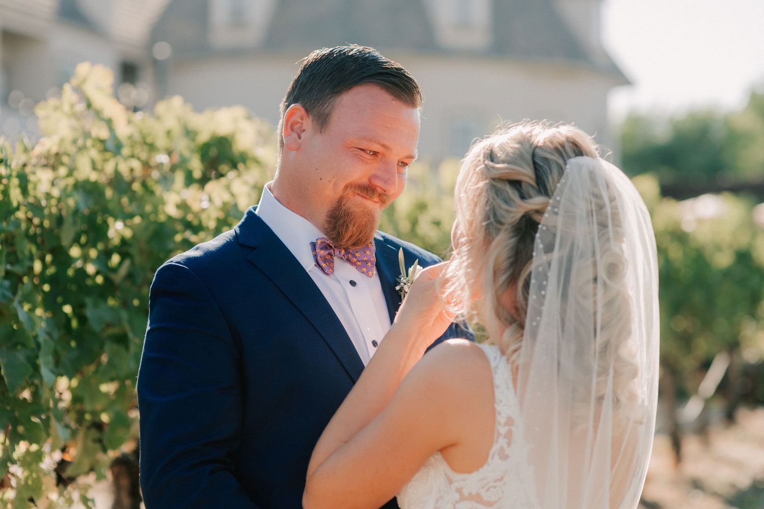 Bride and groom share a tender moment in a sunlit vineyard, groom in navy suit with pink bow tie.