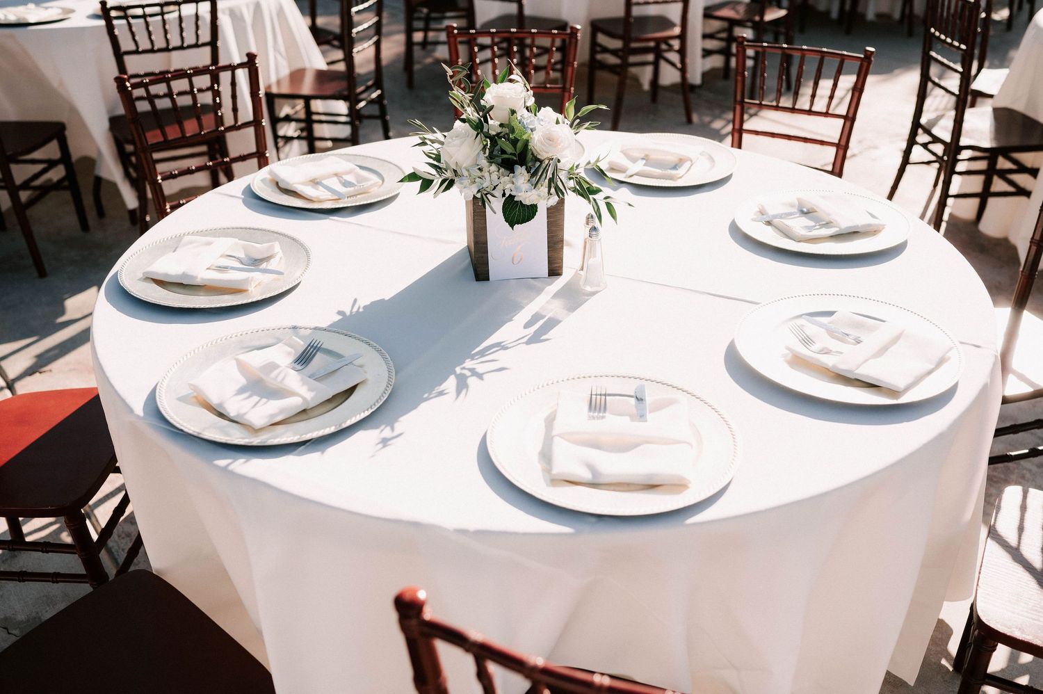 Elegant round wedding reception table with white linen, chiavari chairs, white plates, and a floral centerpiece.
