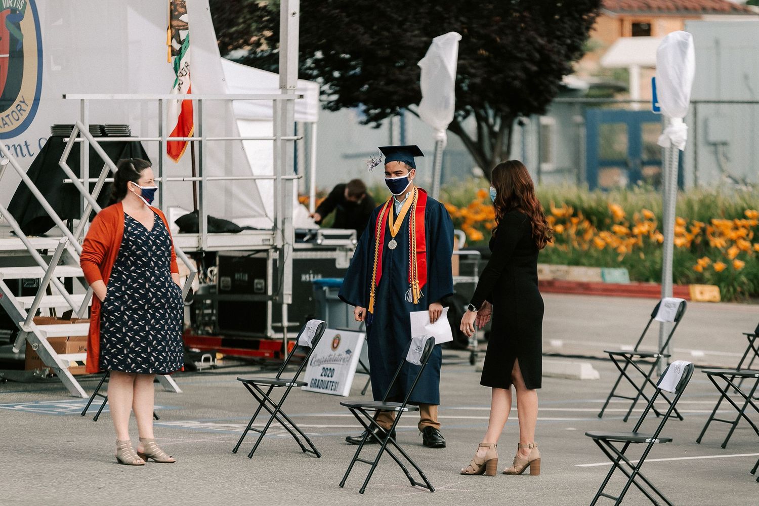 Graduate in cap and gown with mask poses with two women at outdoor socially distanced graduation ceremony.