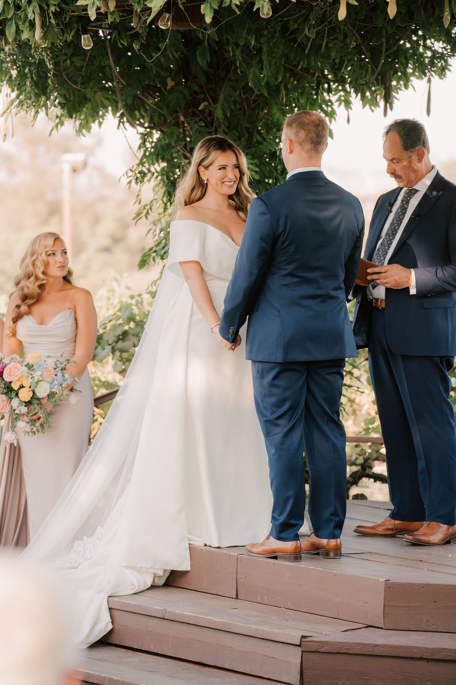 Bride in white off-shoulder gown holds hands with groom in navy suit during outdoor wedding ceremony under lush greenery.